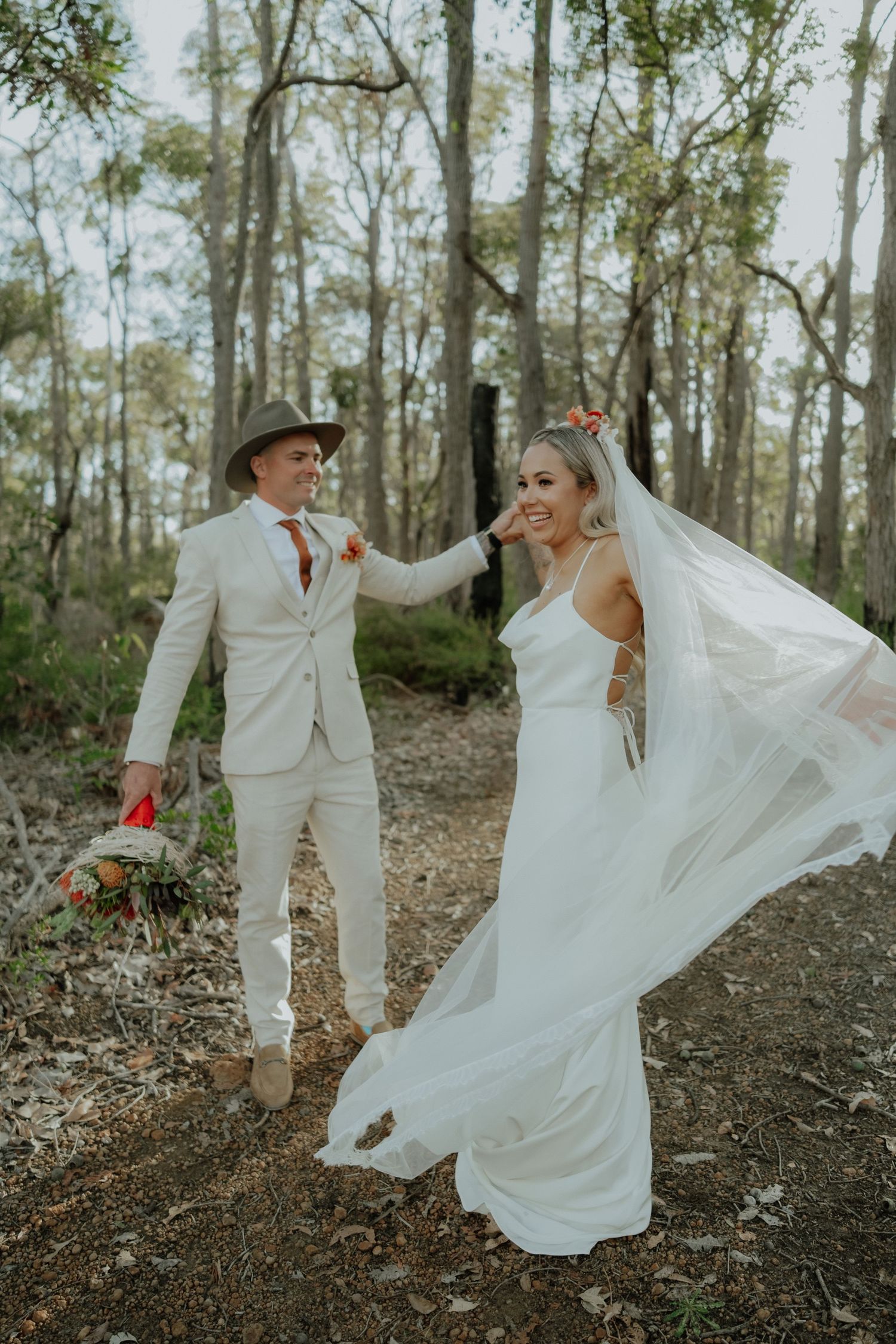 Groom in cream suit and hat twirling bride in flowing white gown through sunlit Australian bushland.