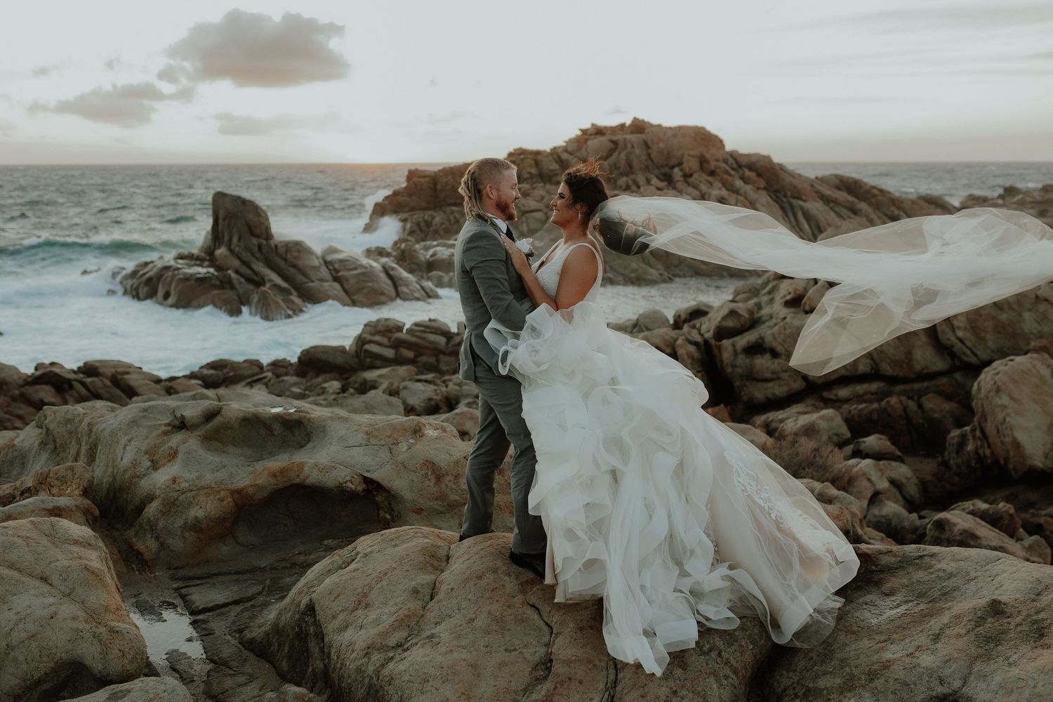 Bride and groom share romantic moment on rocky coastal cliffs at sunset.