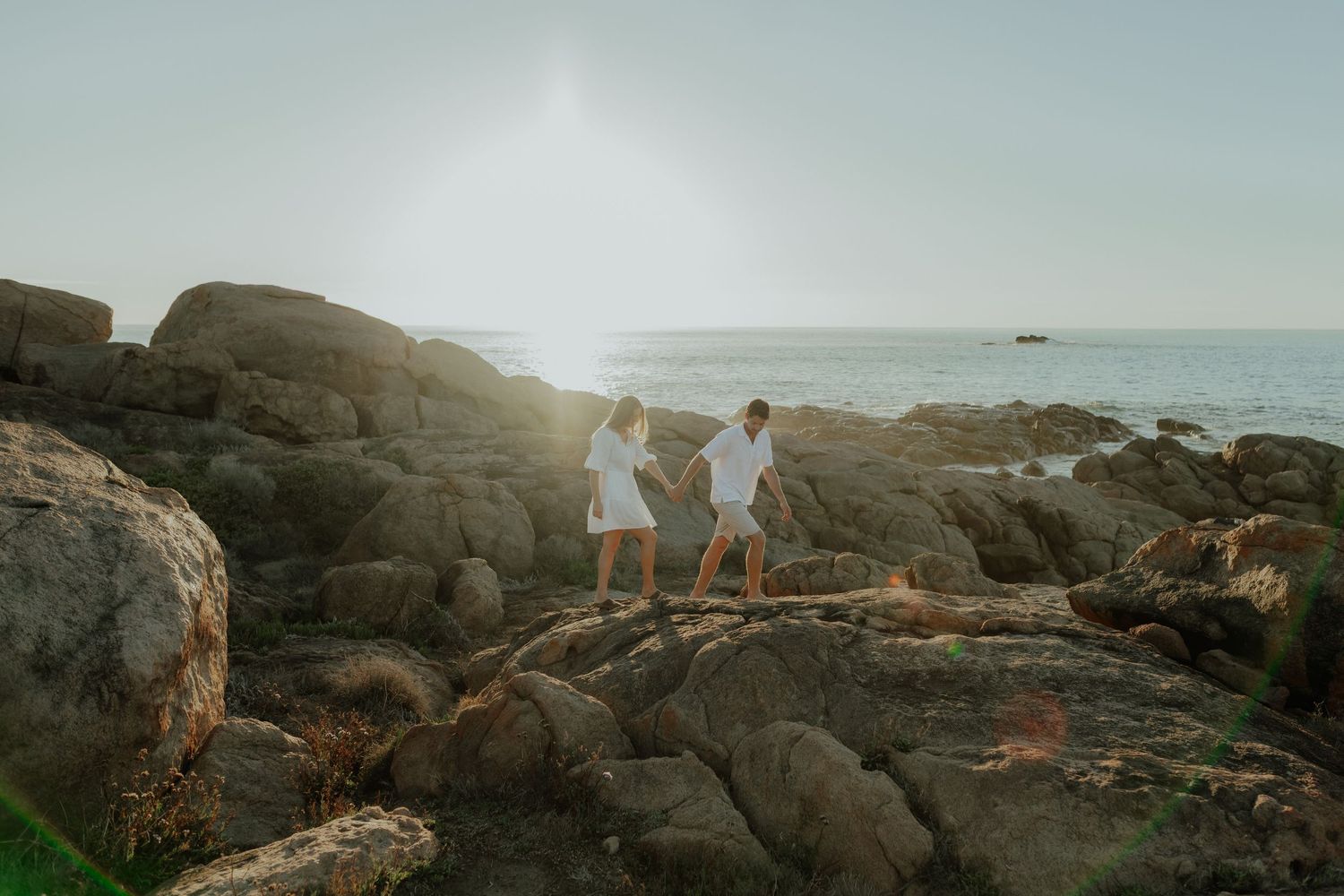Two people in matching white outfits standing on coastal rocks with ocean and sunlight behind them.