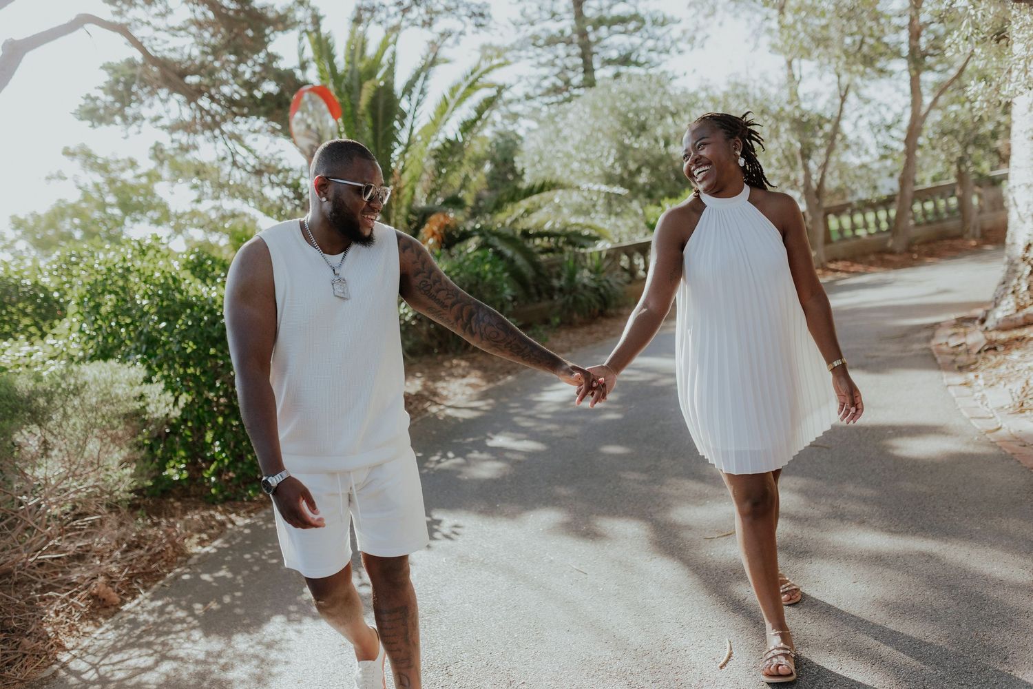 Joyful couple in coordinating white outfits holding hands while strolling along a sunny path.