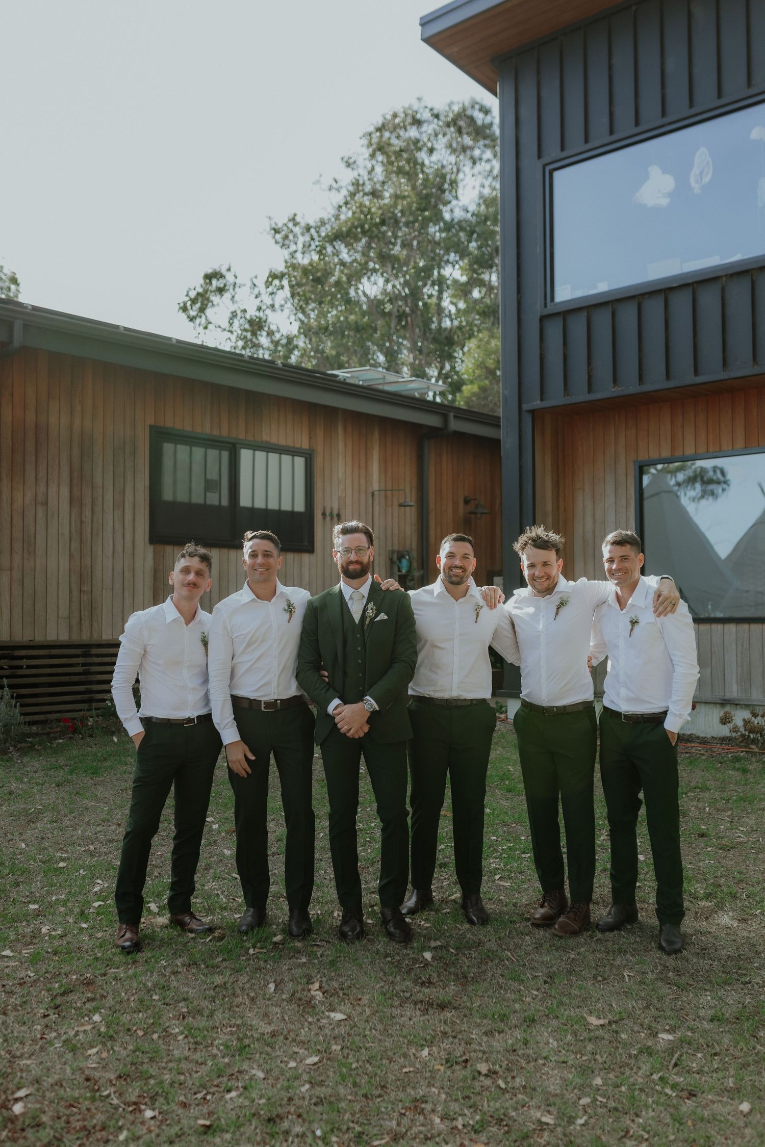 Groom in dark suit stands with five groomsmen in white shirts outside modern timber and dark clad building.