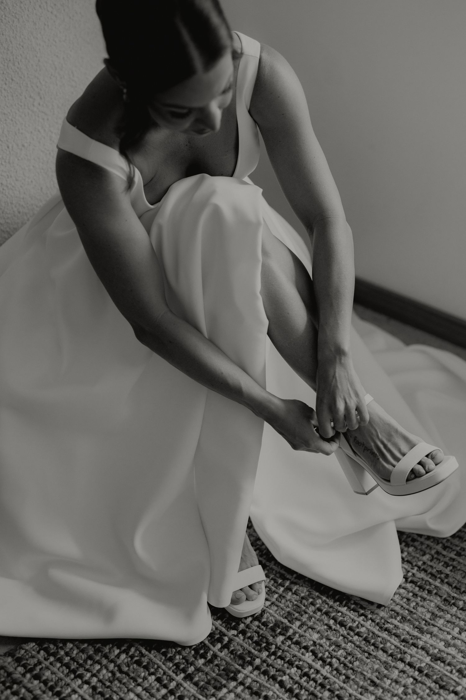 Black and white photo of bride in wedding dress fastening white heeled sandals while seated.