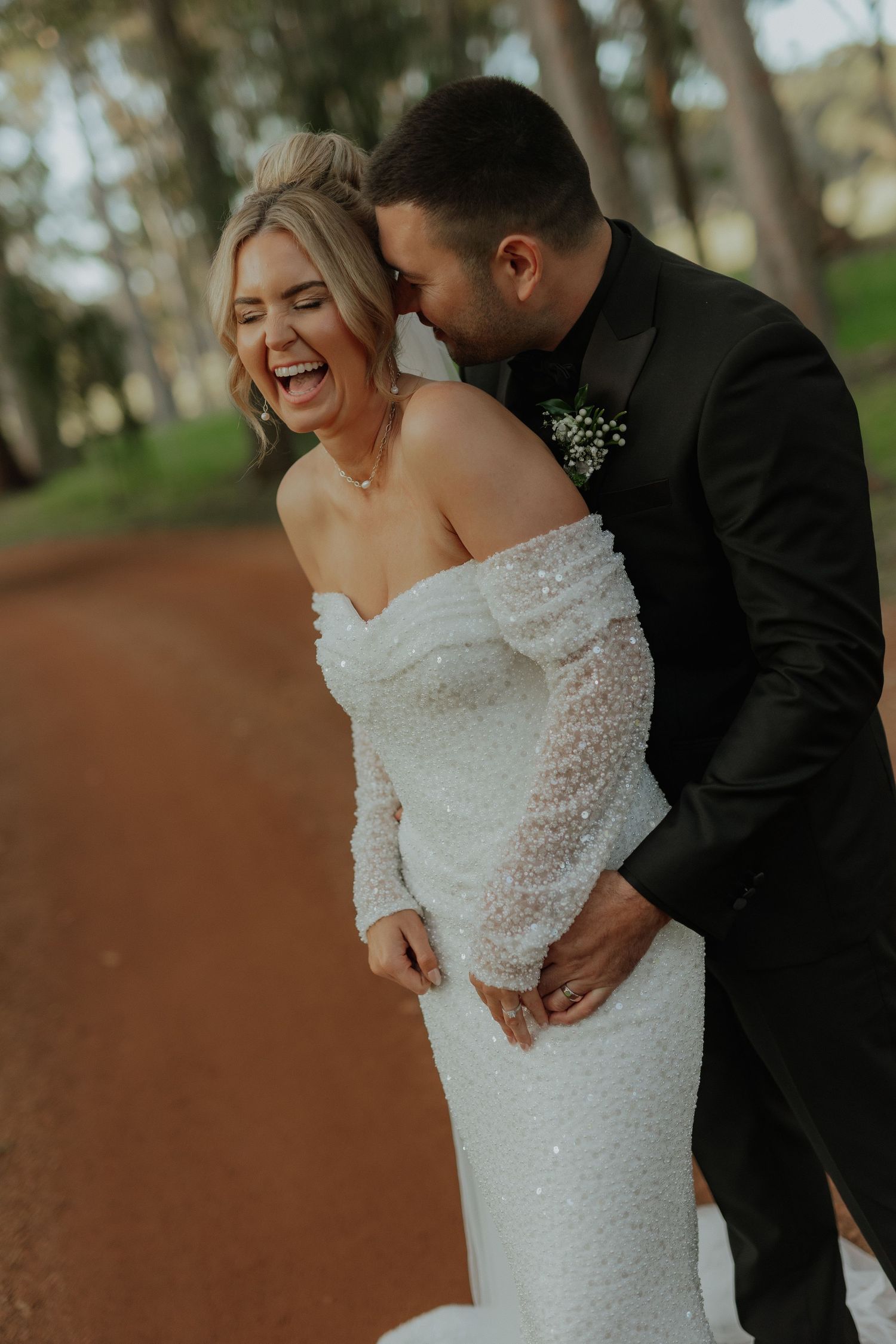 Laughing bride in beaded off-shoulder gown embraced by groom in black suit on dirt path.