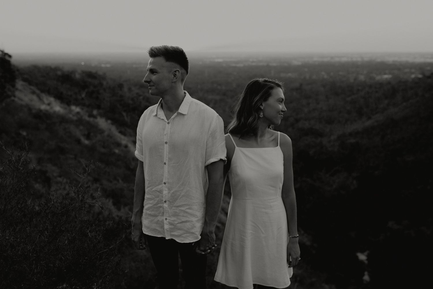 Black and white photo of a couple holding hands on a hilltop overlooking a scenic landscape.