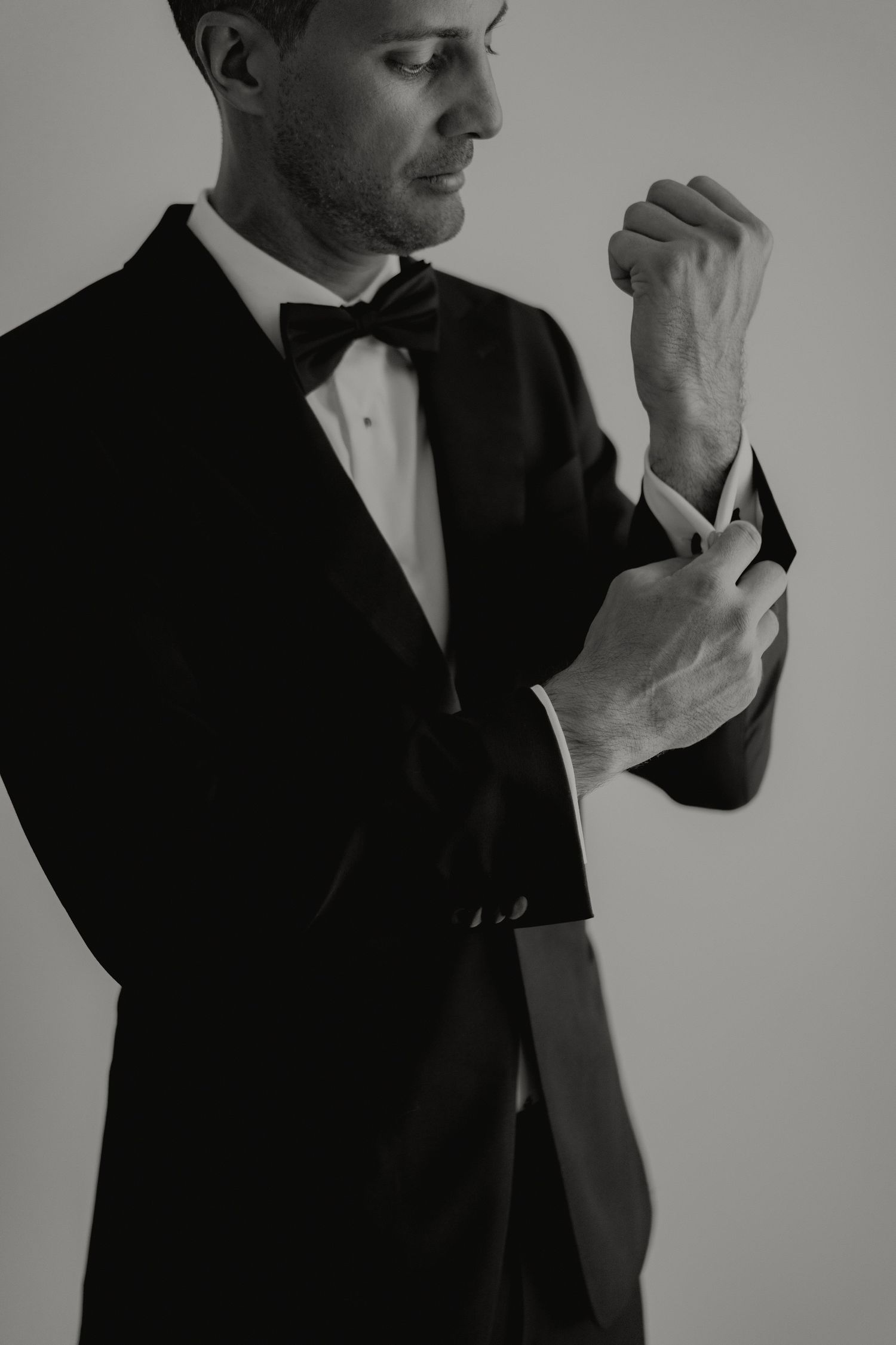 Groom in black tuxedo adjusting cufflinks, elegant black and white wedding portrait.
