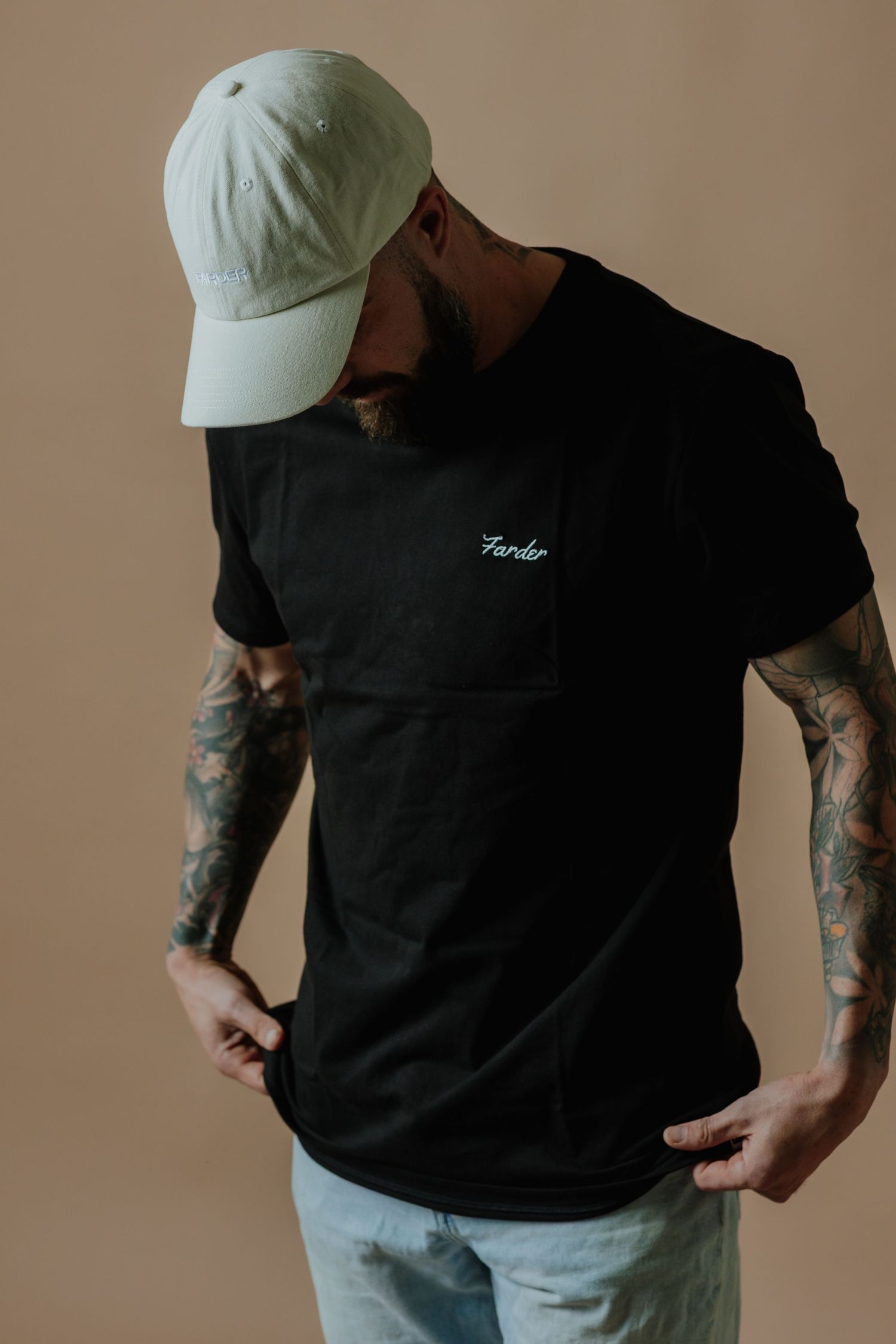 Tattooed bearded man wearing a black Farder t-shirt and white cap posed against a beige background.