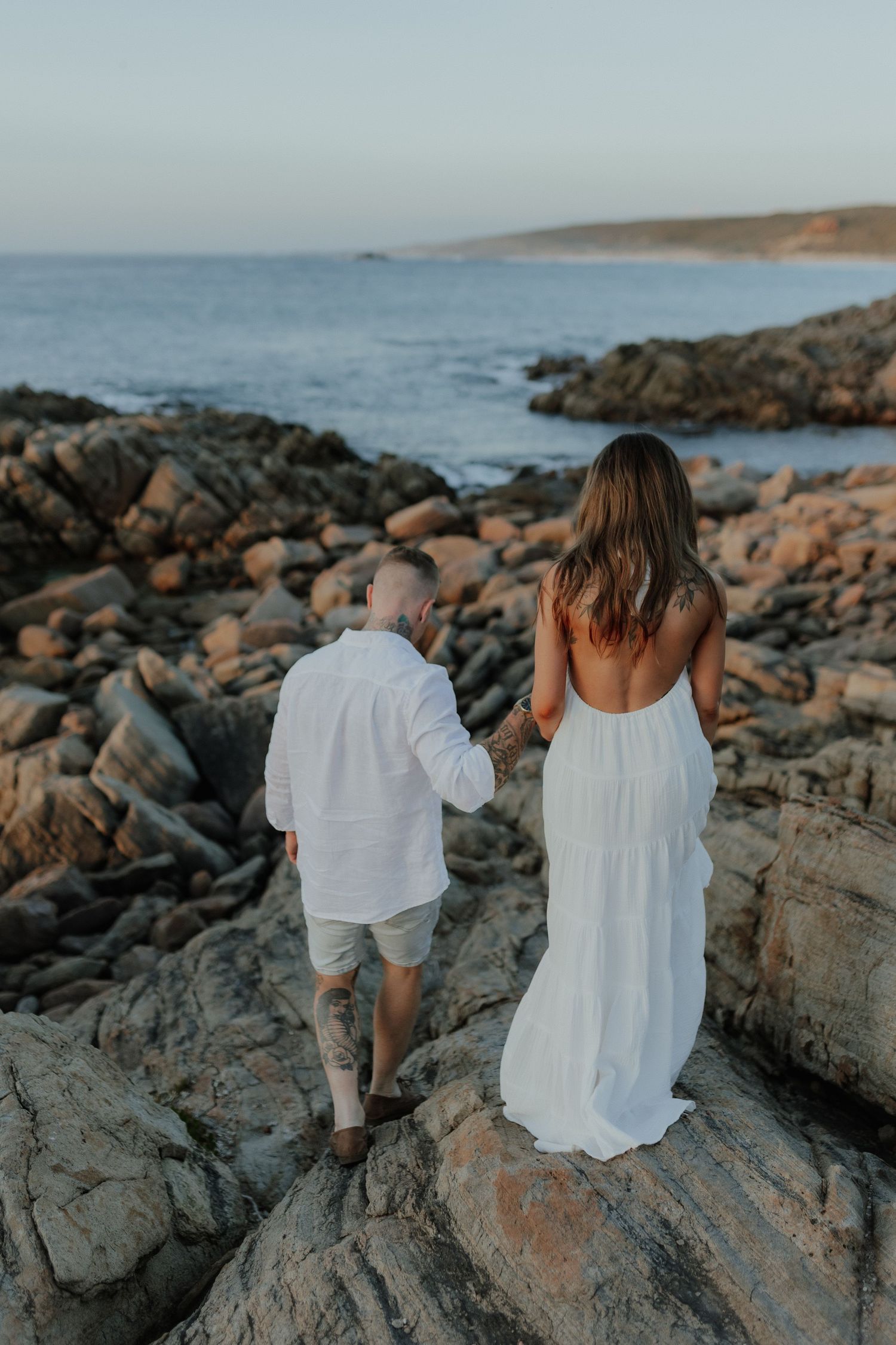 Couple holding hands walking on rocky coastal shoreline at sunset, woman in white backless dress.