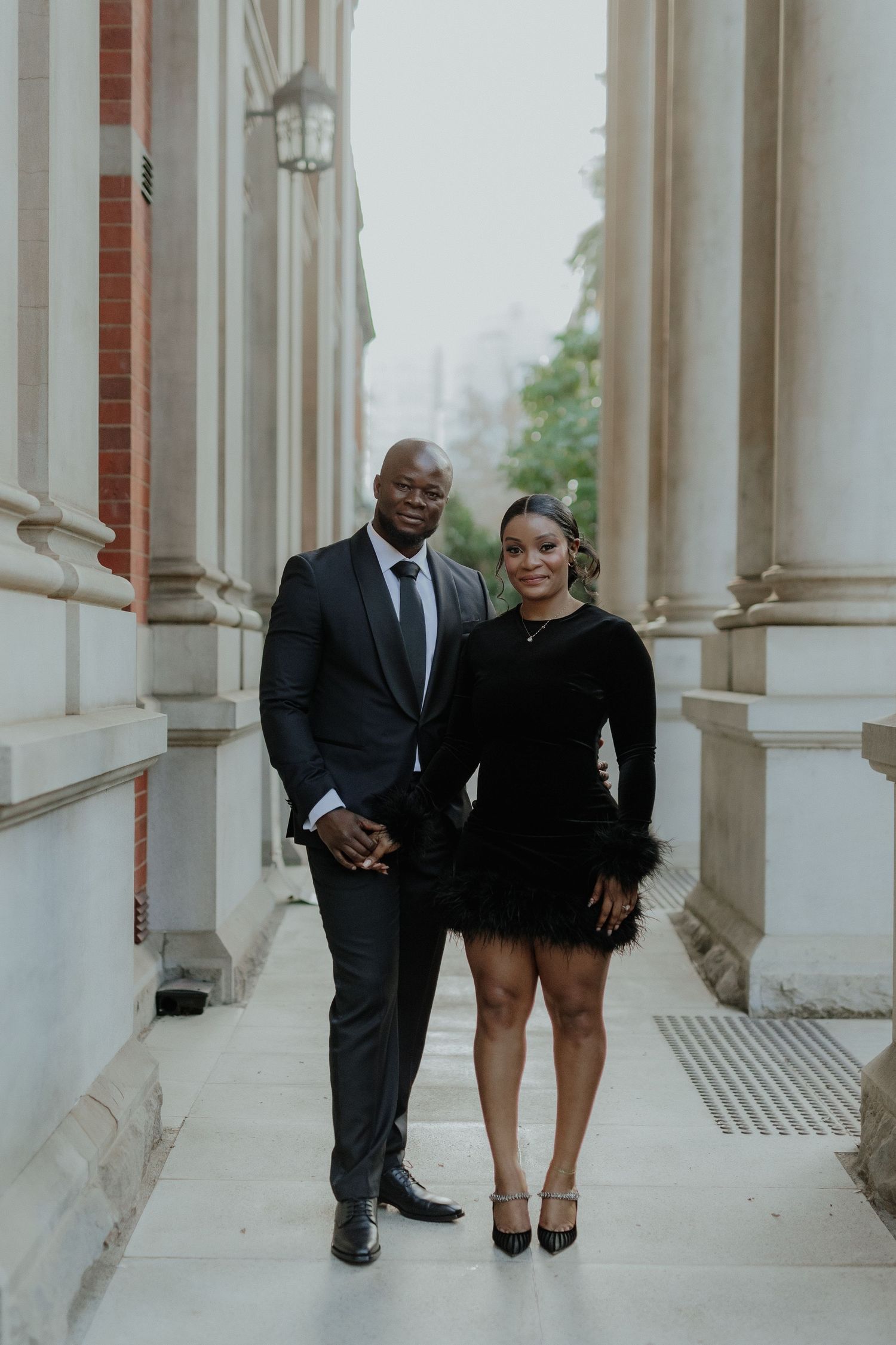 Stylish couple in matching black outfits posing between classical stone columns outdoors.