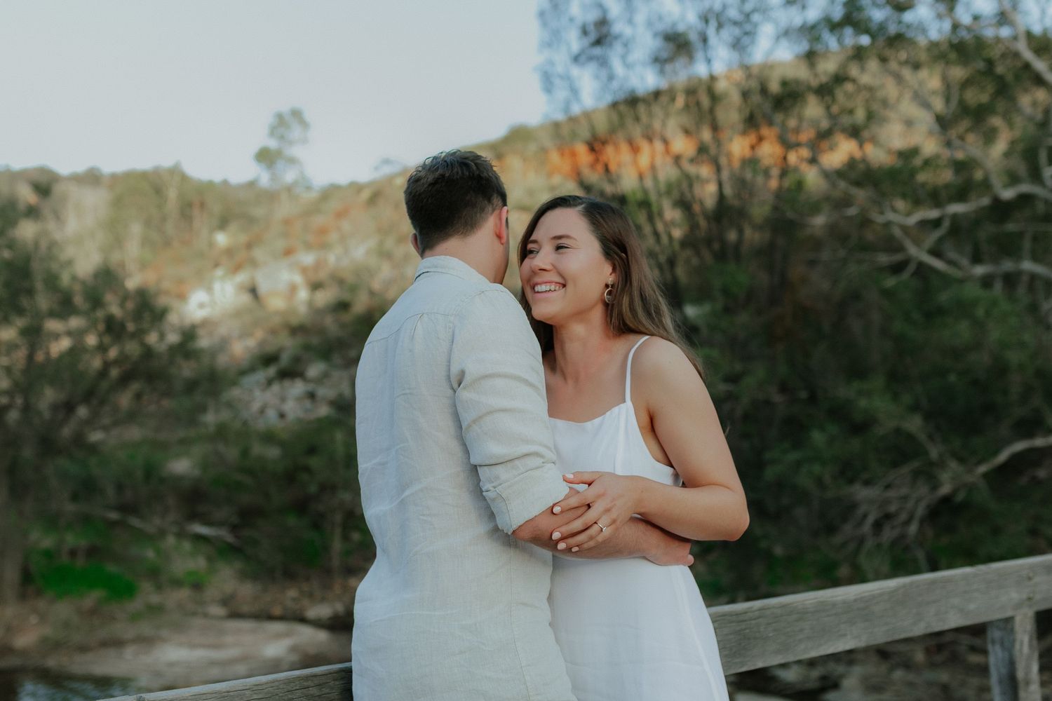 Smiling woman embraces her partner outdoors with a scenic hillside backdrop during golden hour.