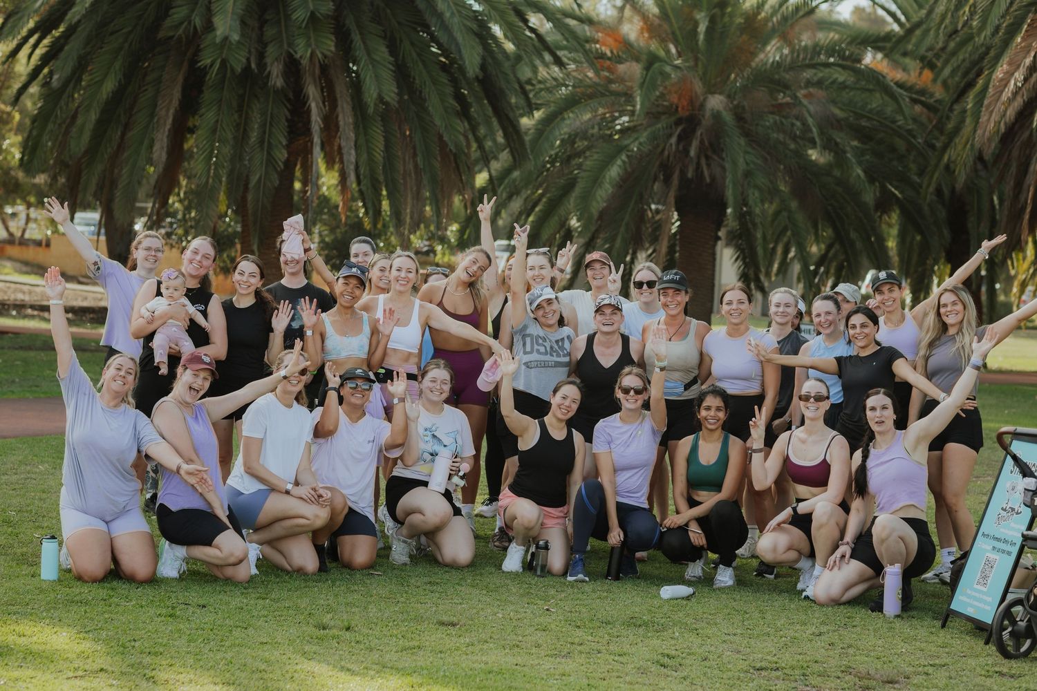 Large group of women in activewear posing happily in a park surrounded by palm trees.