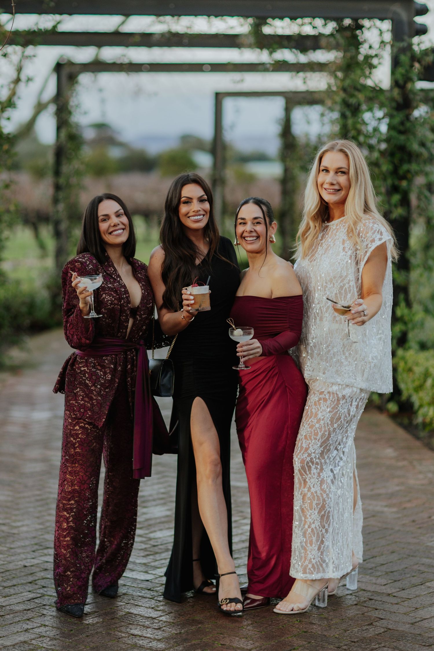 Four women in elegant formal gowns posing together at an outdoor winery event.