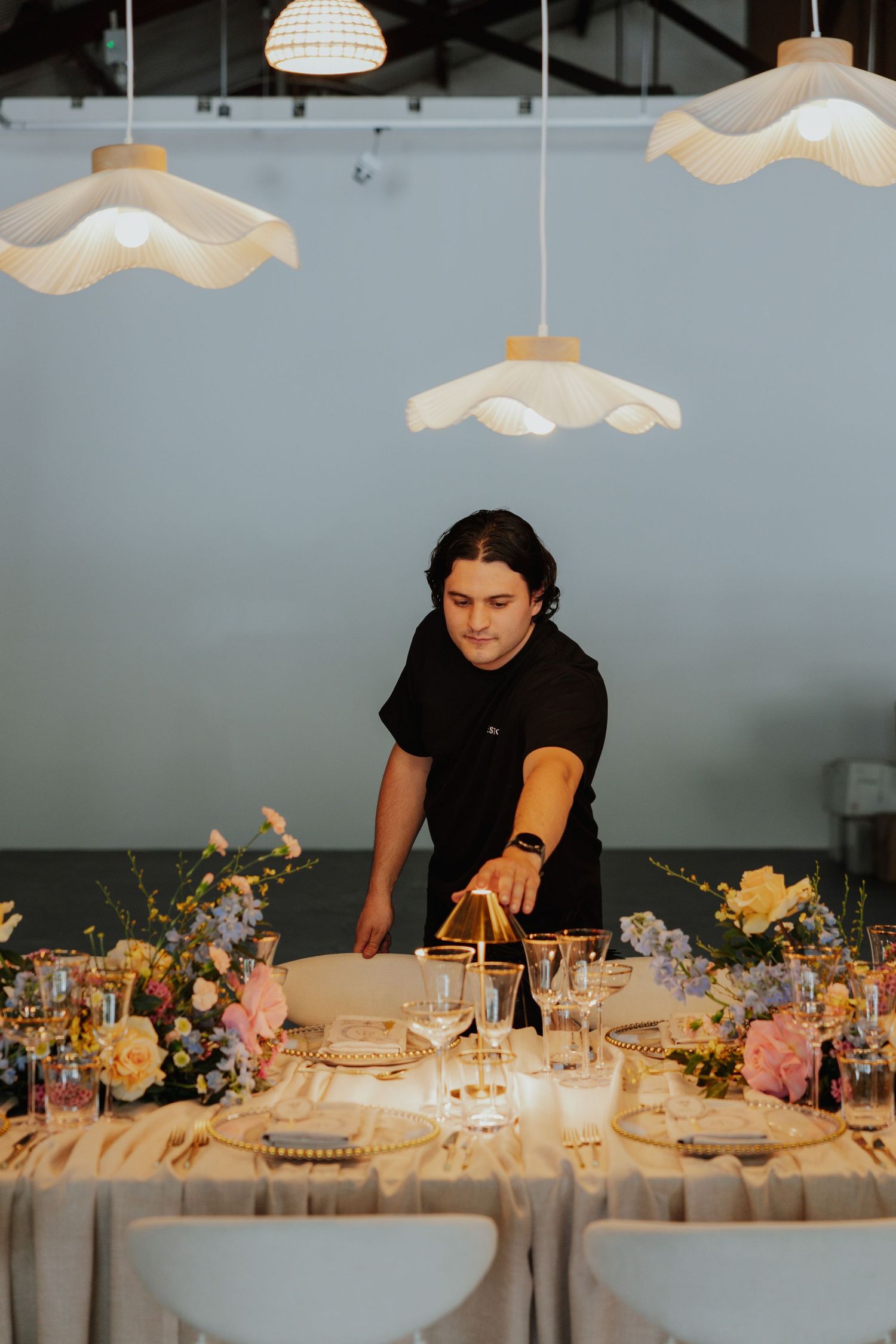 A server arranges place settings on an elegantly decorated wedding reception table with floral centerpieces and pendant lights.