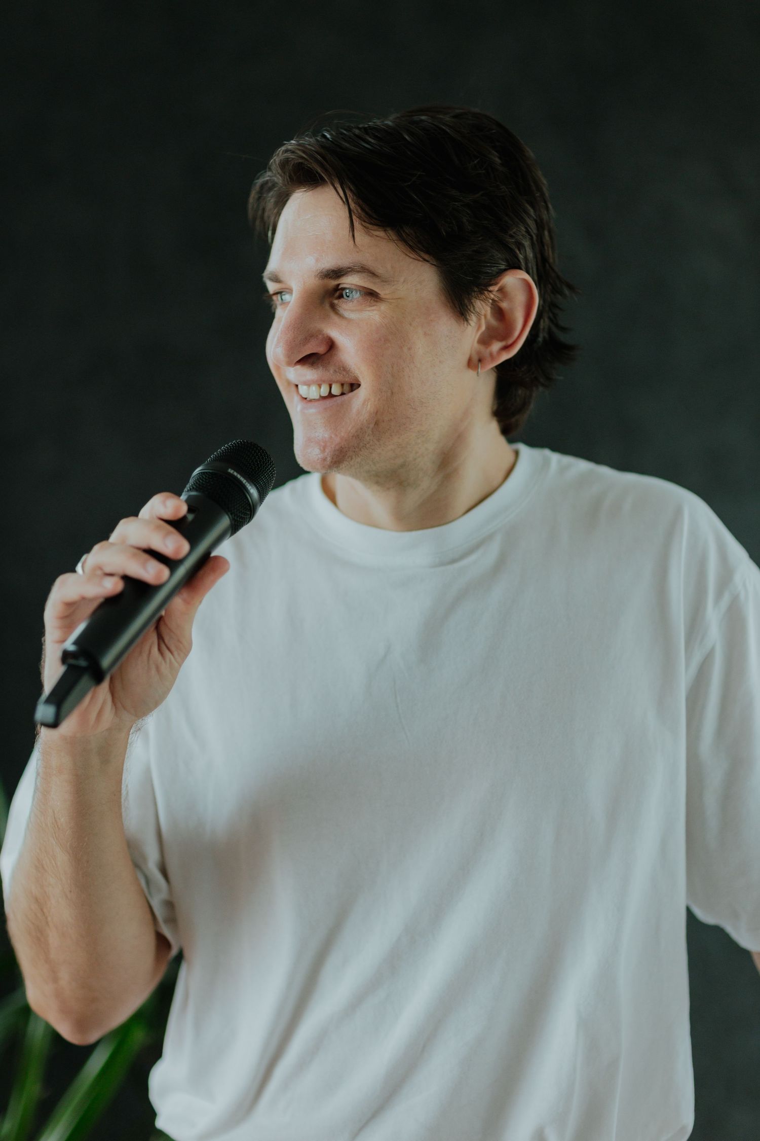 Smiling man in white t-shirt holding a black microphone against dark background.