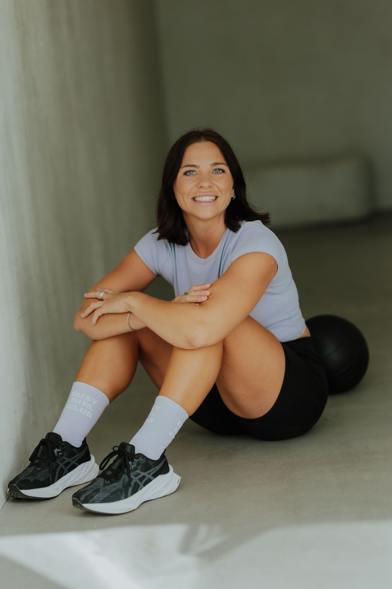 Smiling woman in white t-shirt sitting on floor with black exercise ball behind her.