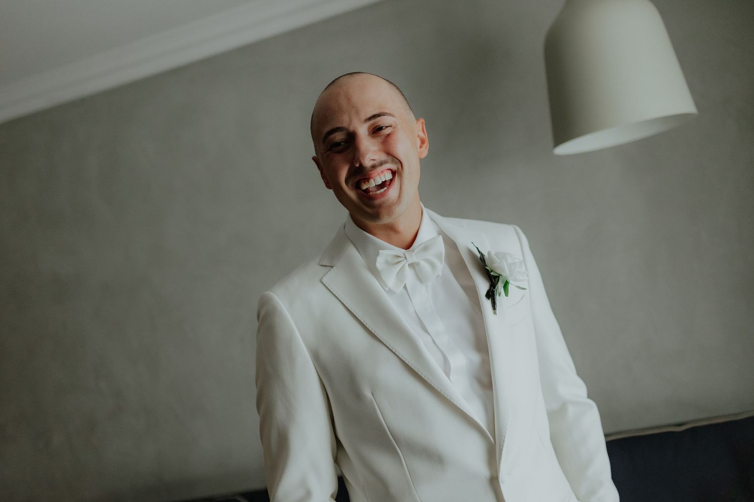 Smiling bald groom in white tuxedo with boutonniere on wedding day.
