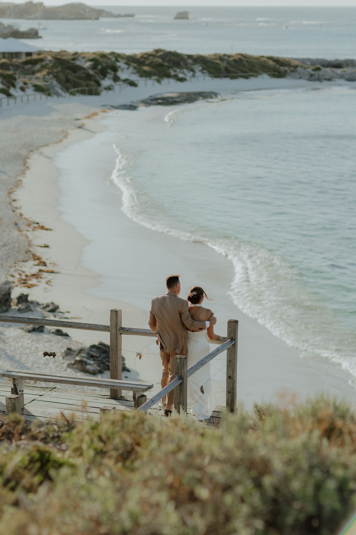 Bride and groom stand at wooden railing overlooking stunning white sand beach at sunset.