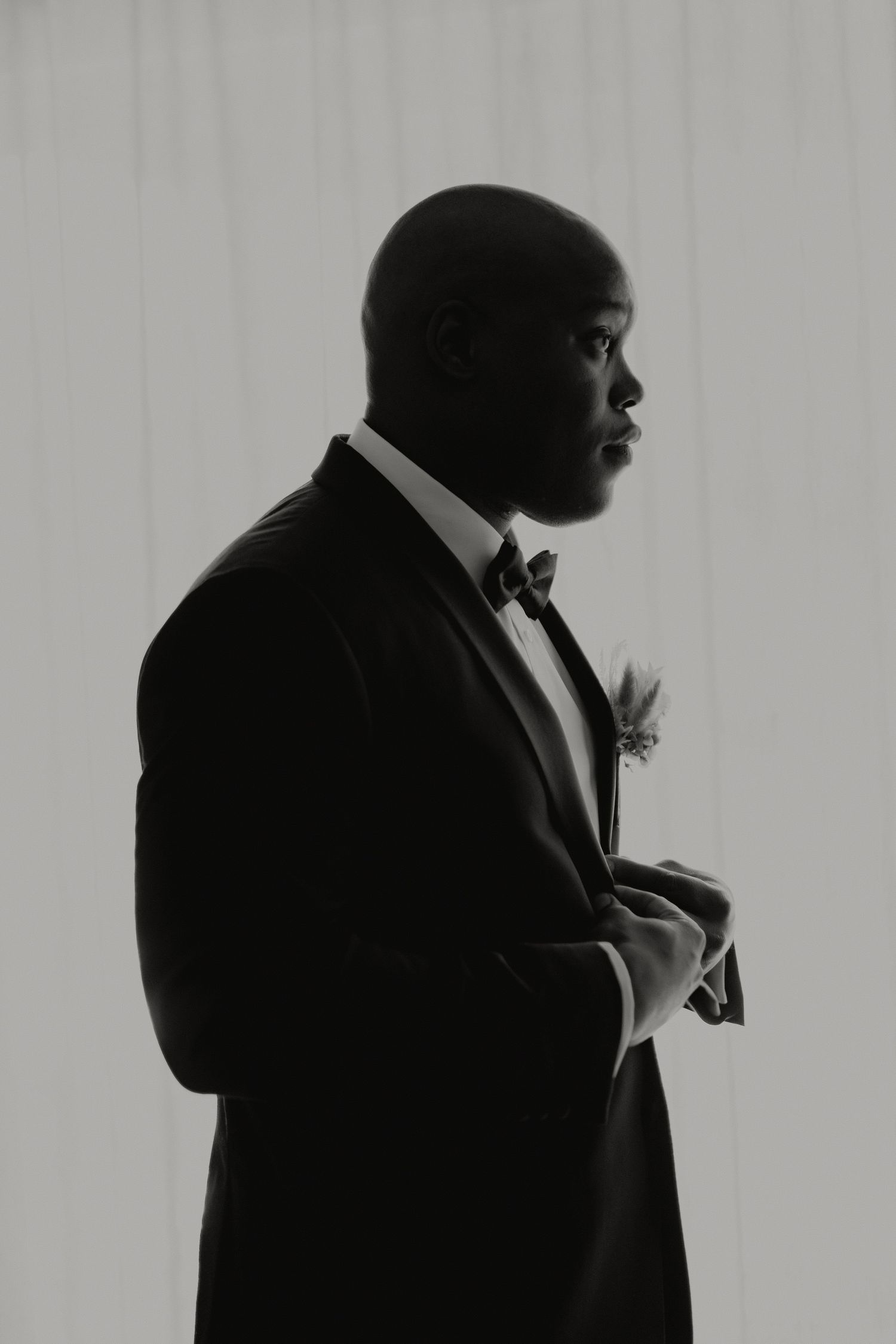 Dramatic black and white silhouette portrait of groom in tuxedo with boutonniere adjusting cuffs.