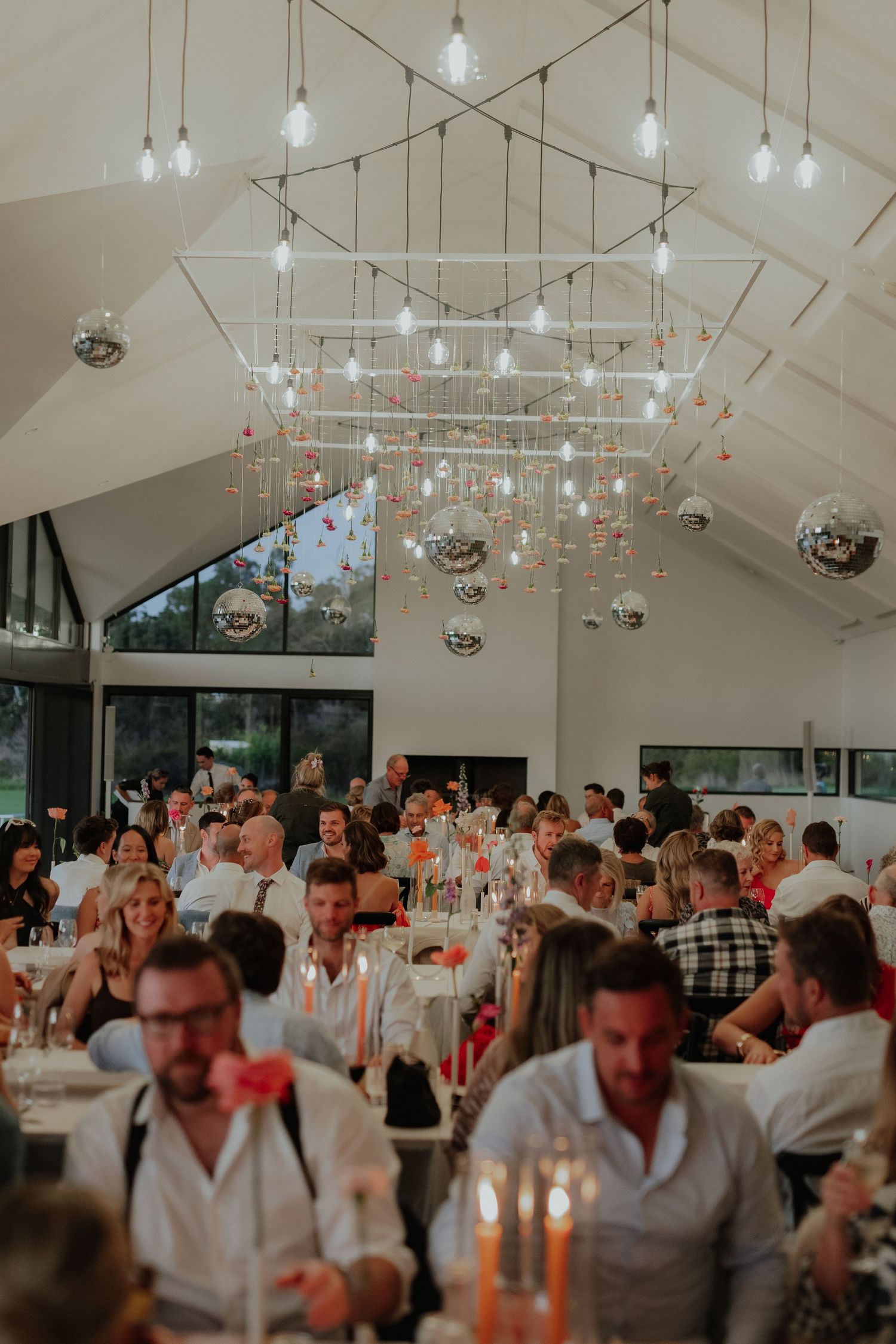 Elegant wedding reception hall with Edison bulb string lights, disco balls, and guests seated at decorated tables.