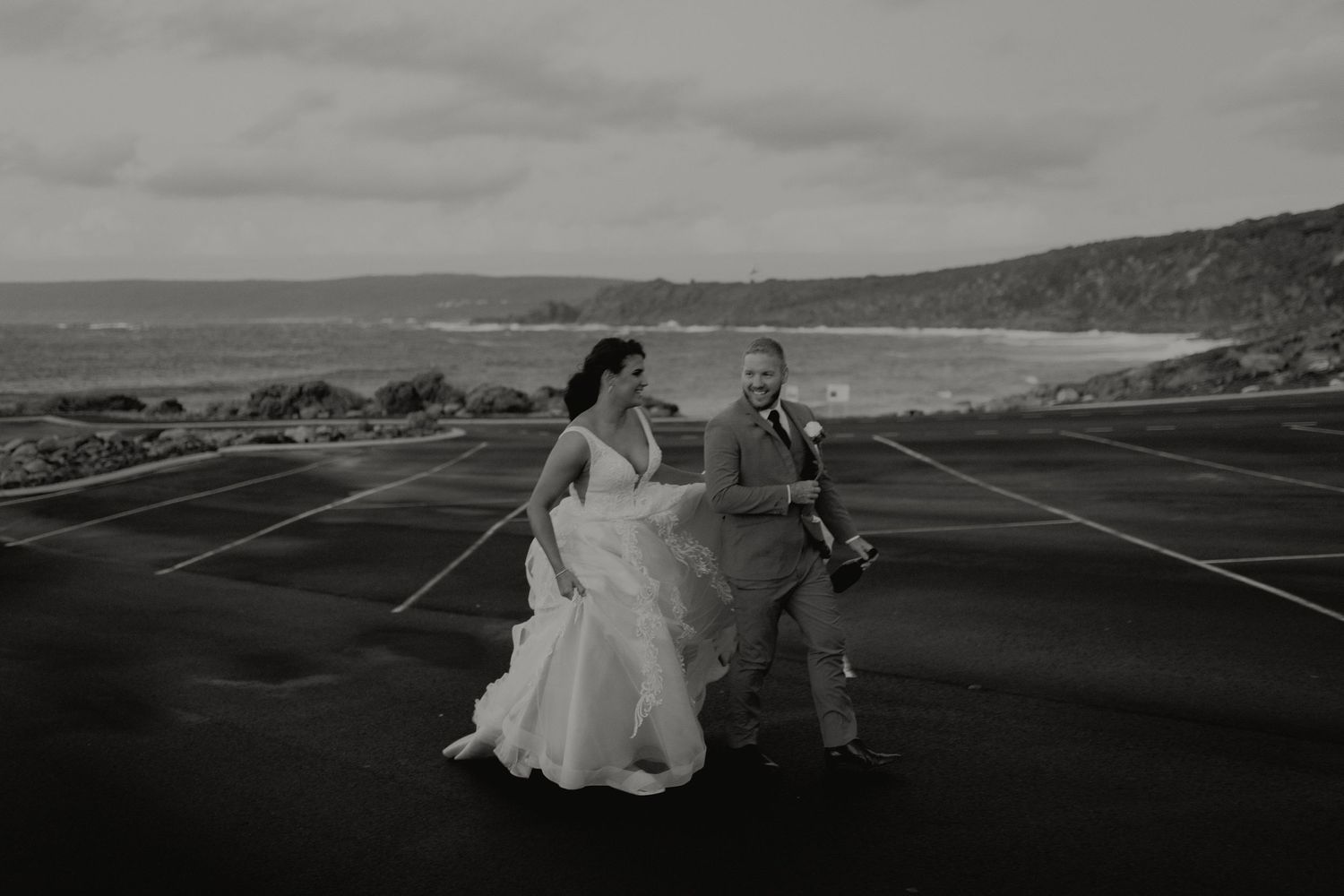 Bride and groom walk hand in hand across coastal parking lot in black and white photo.