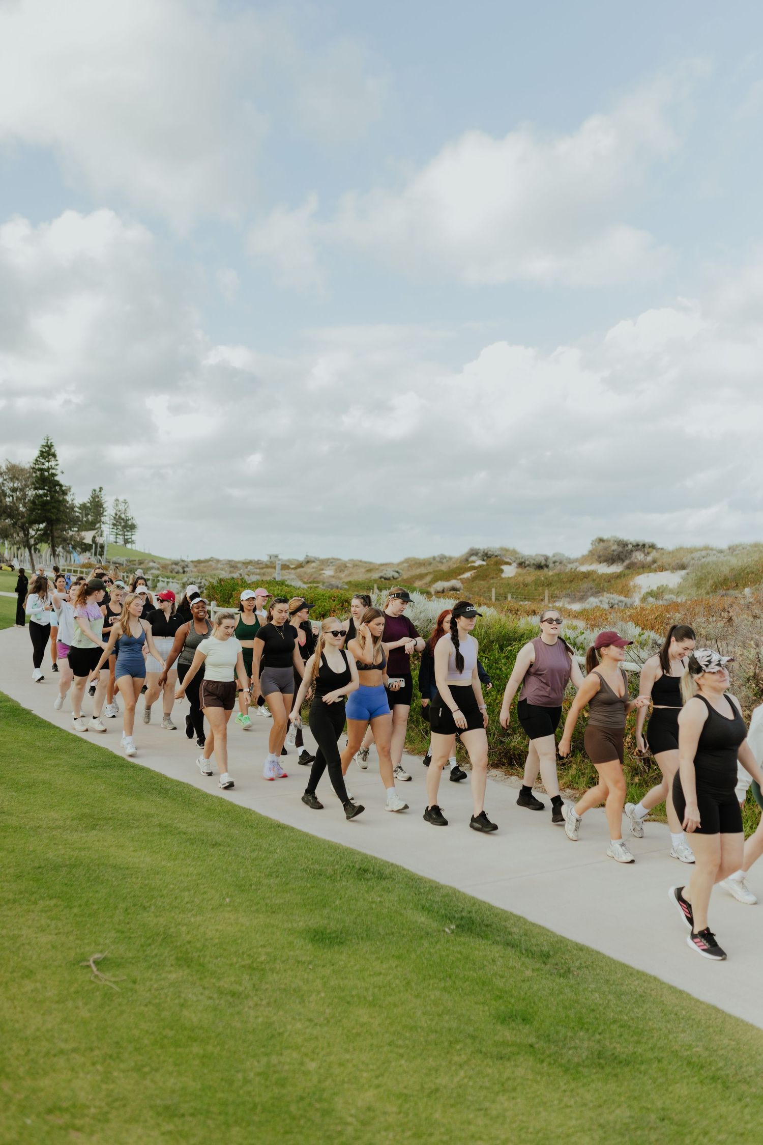 Large group of women in athletic wear walking along a coastal pathway on a cloudy day.