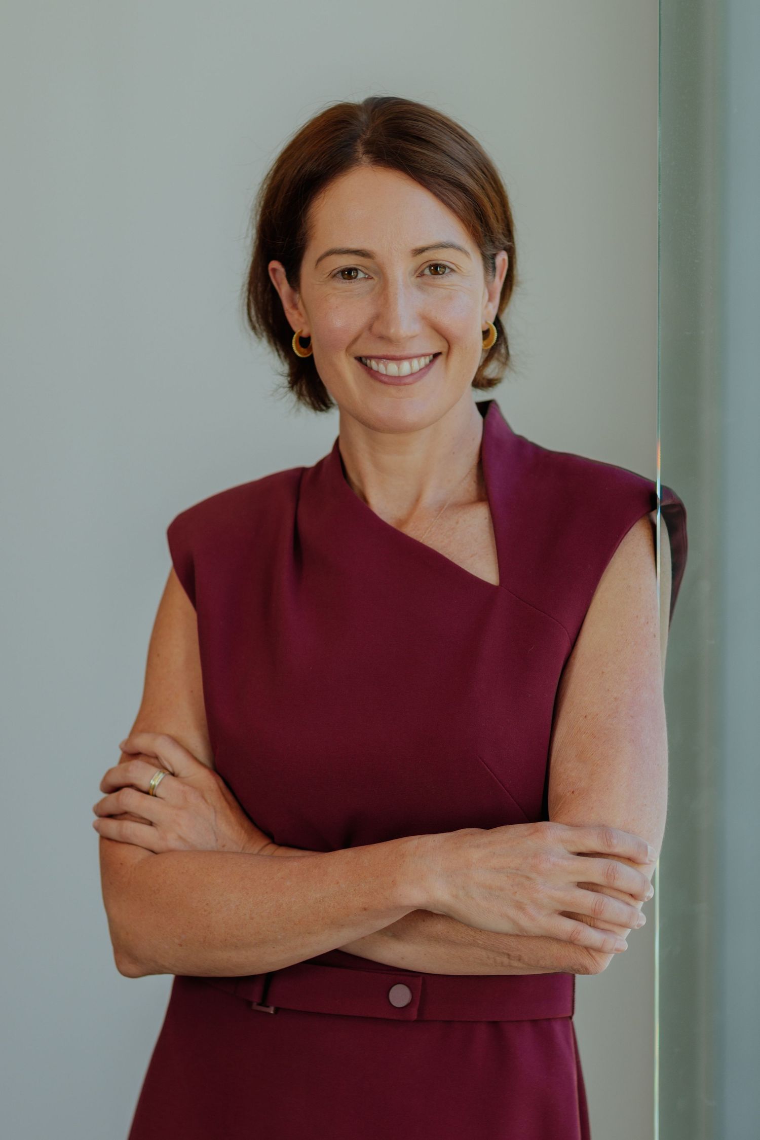 Confident professional woman in burgundy dress standing with arms crossed smiling in a modern office.