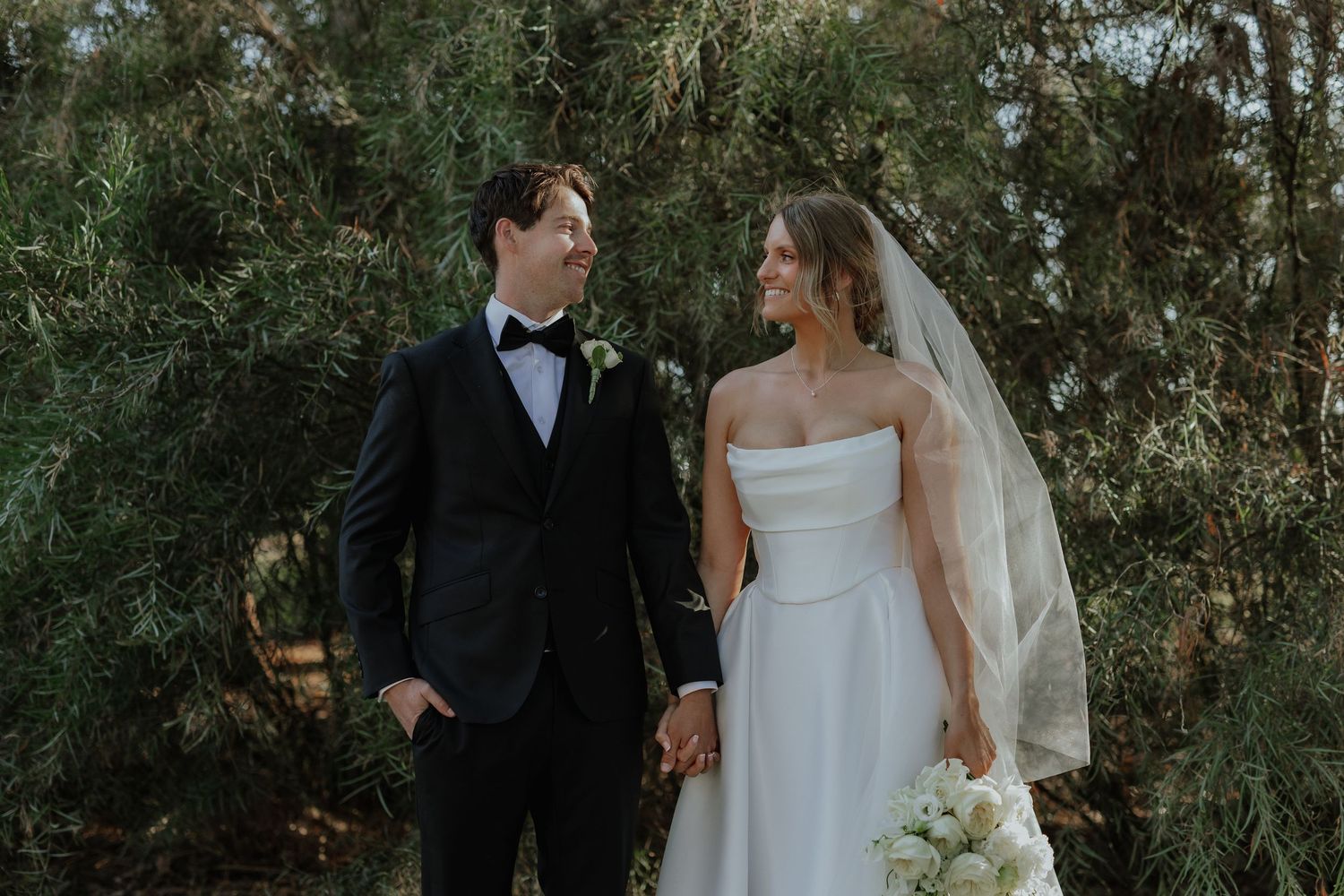 Bride in strapless white gown and groom in black tuxedo holding hands among green trees.