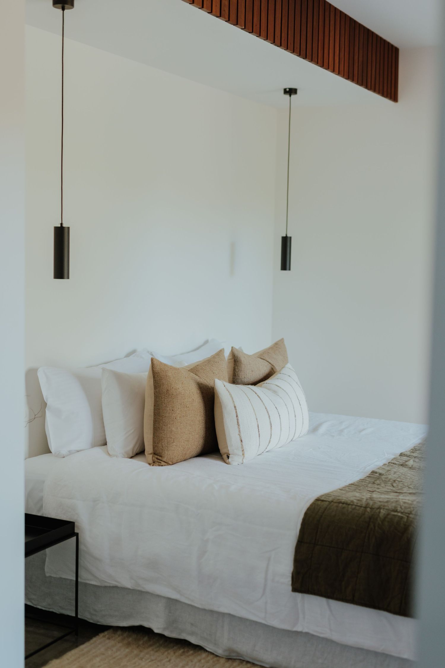 Elegant bedroom with white linen, tan striped cushions, olive throw and twin black cylinder pendant lights.