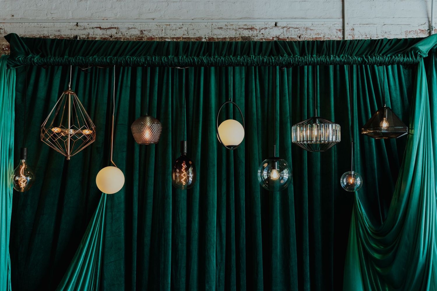 Assorted decorative pendant lights including geometric and globe styles against rich green velvet curtains.