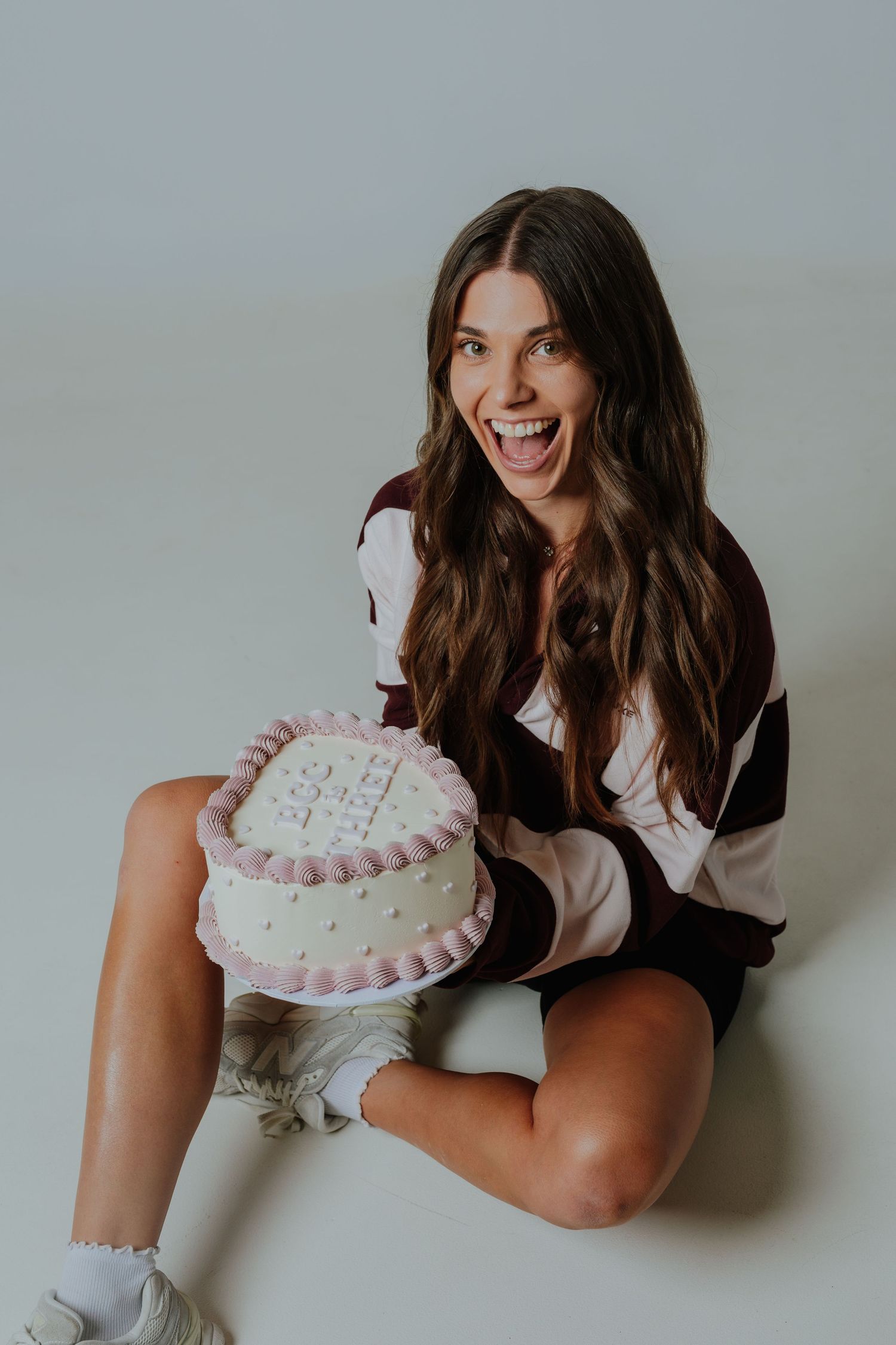 Excited woman sitting on floor holding a pink decorated birthday cake, laughing joyfully.