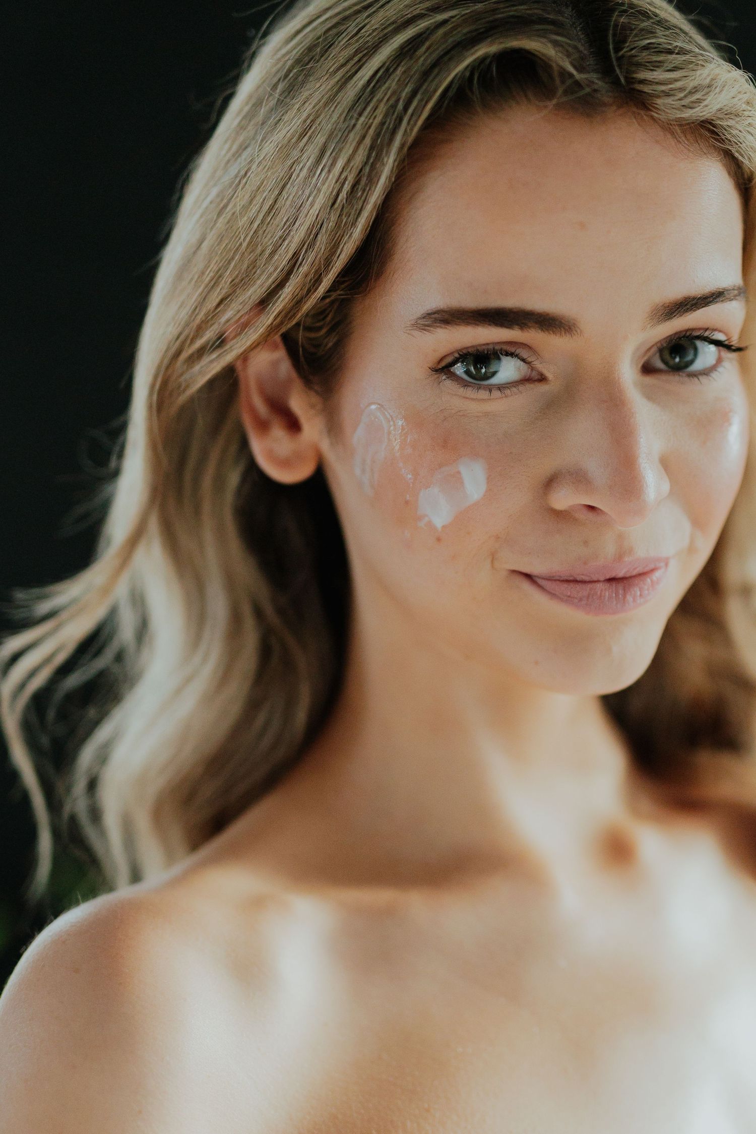 Blonde woman smiling with white moisturizer dots on her cheek demonstrating a skincare application.