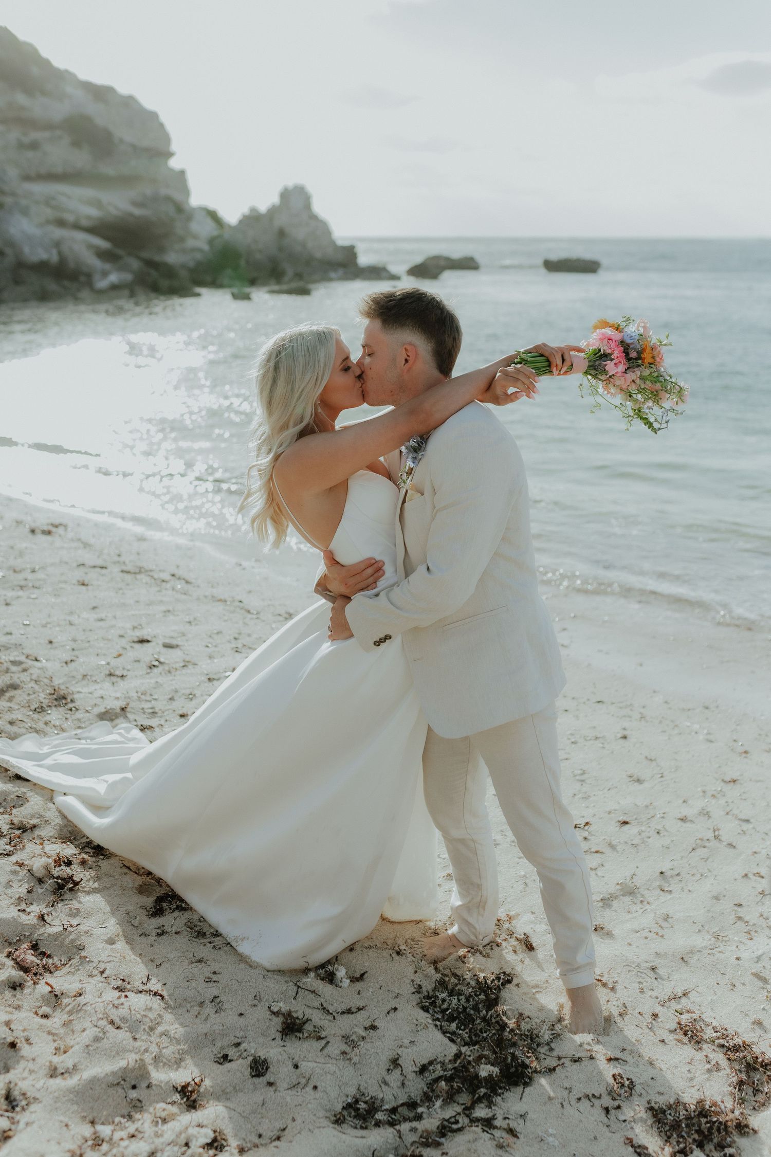 Bride and groom share a romantic kiss on a white sand beach holding a colorful bouquet.
