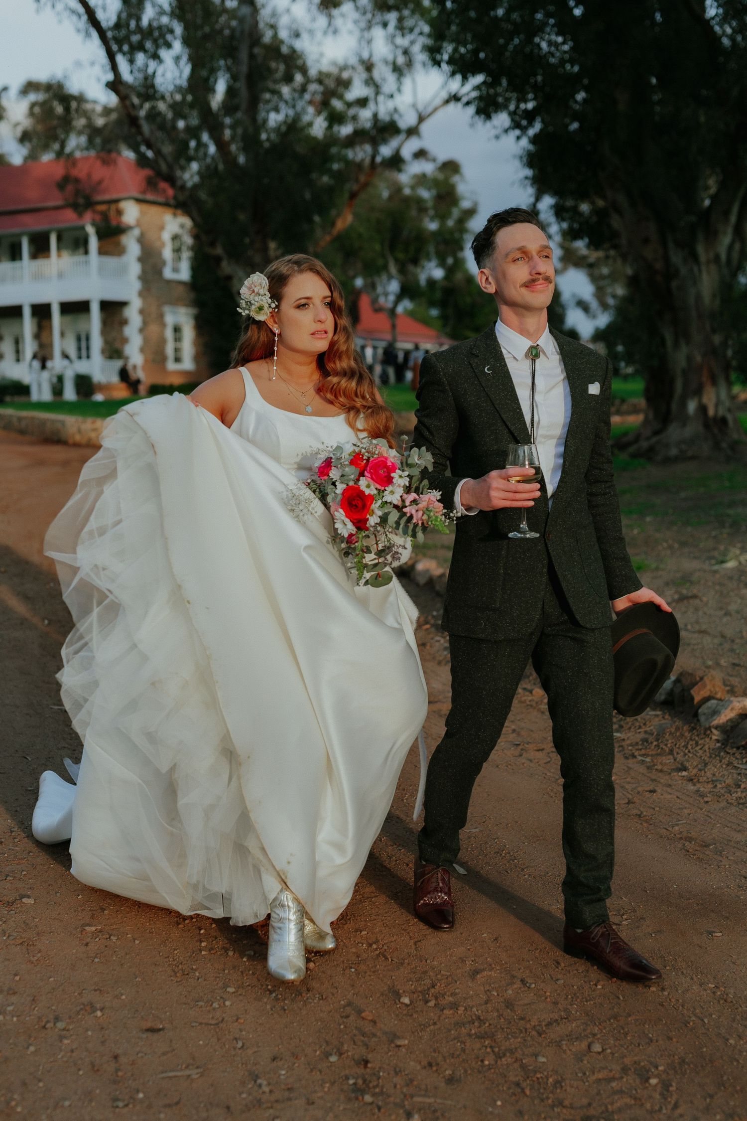 Bride and groom walk together at sunset outside historic estate venue holding bouquet and champagne.
