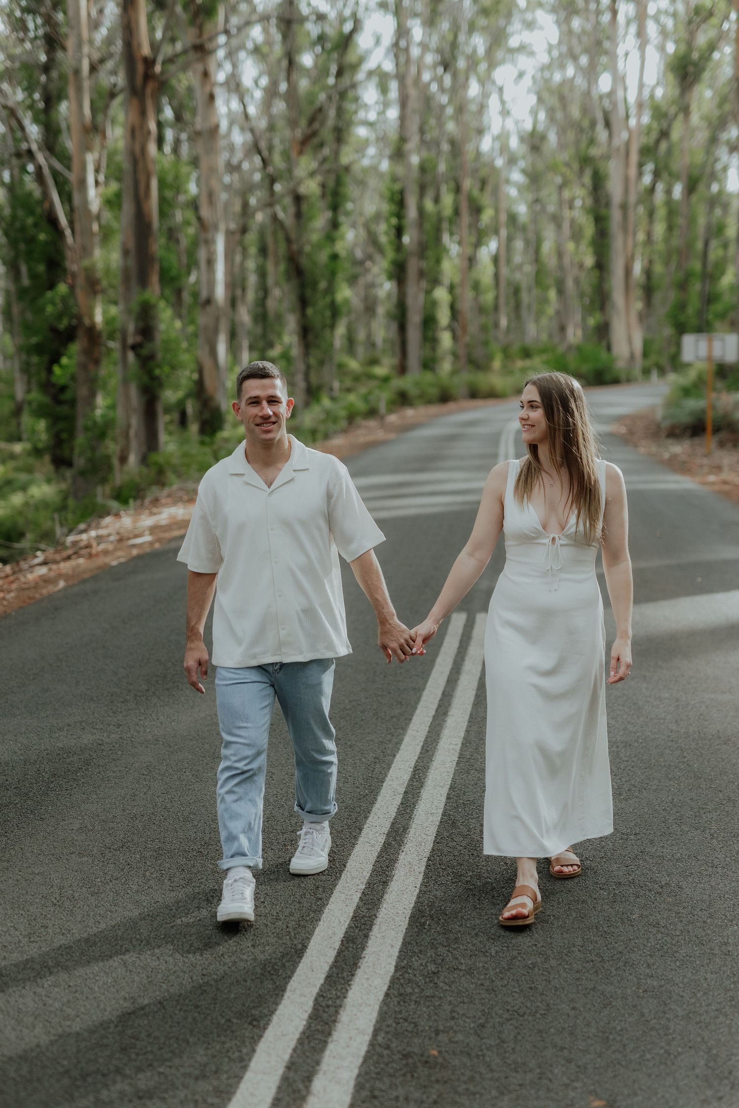 Happy couple in matching white outfits holding hands while walking along a tree-lined road.