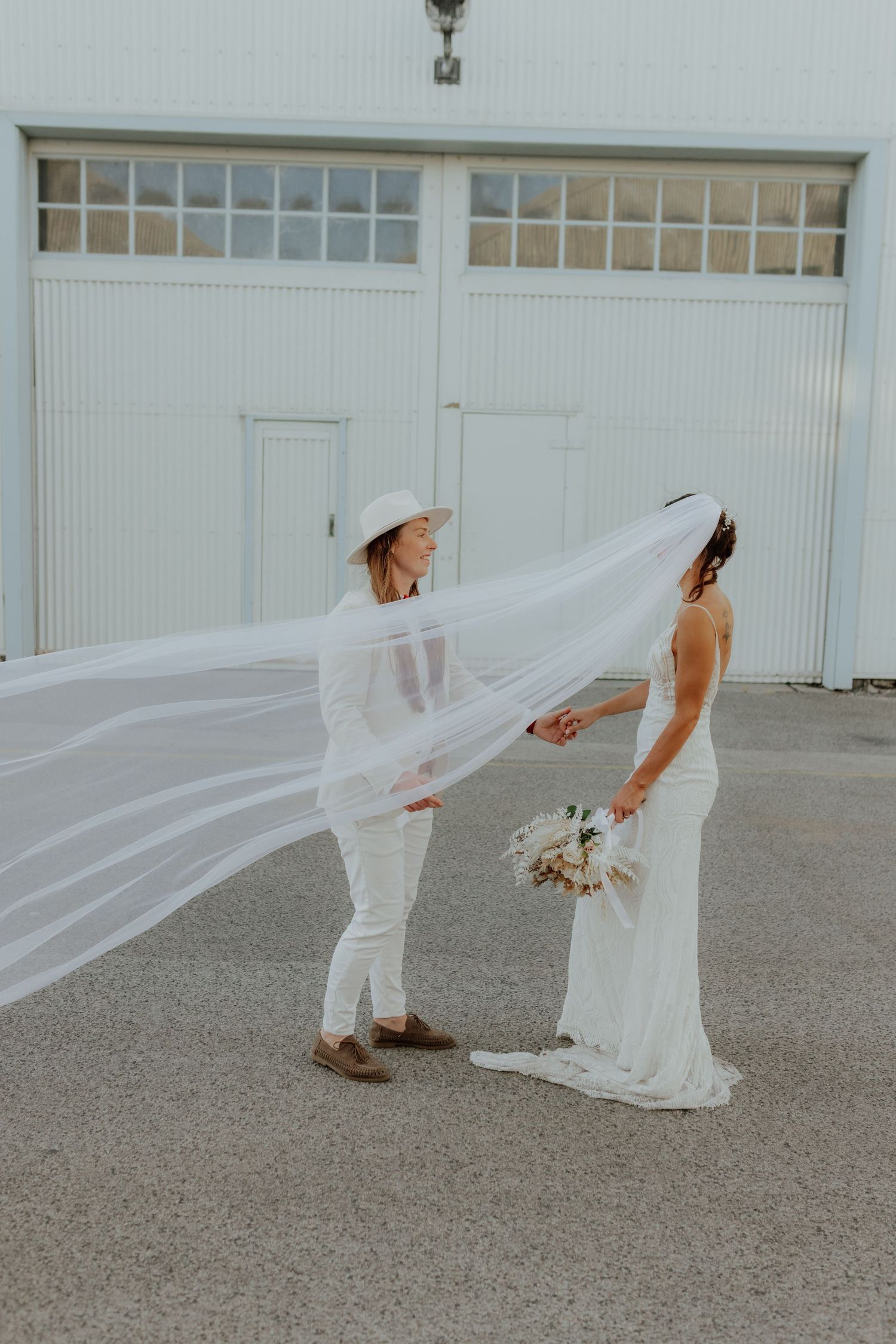 Two brides in white hold hands as long veil billows in wind outside white barn venue.