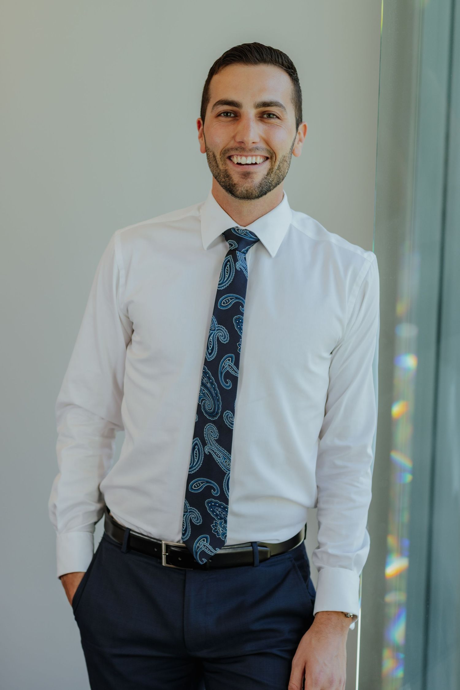 Smiling professional man in white shirt and paisley tie standing in bright office.