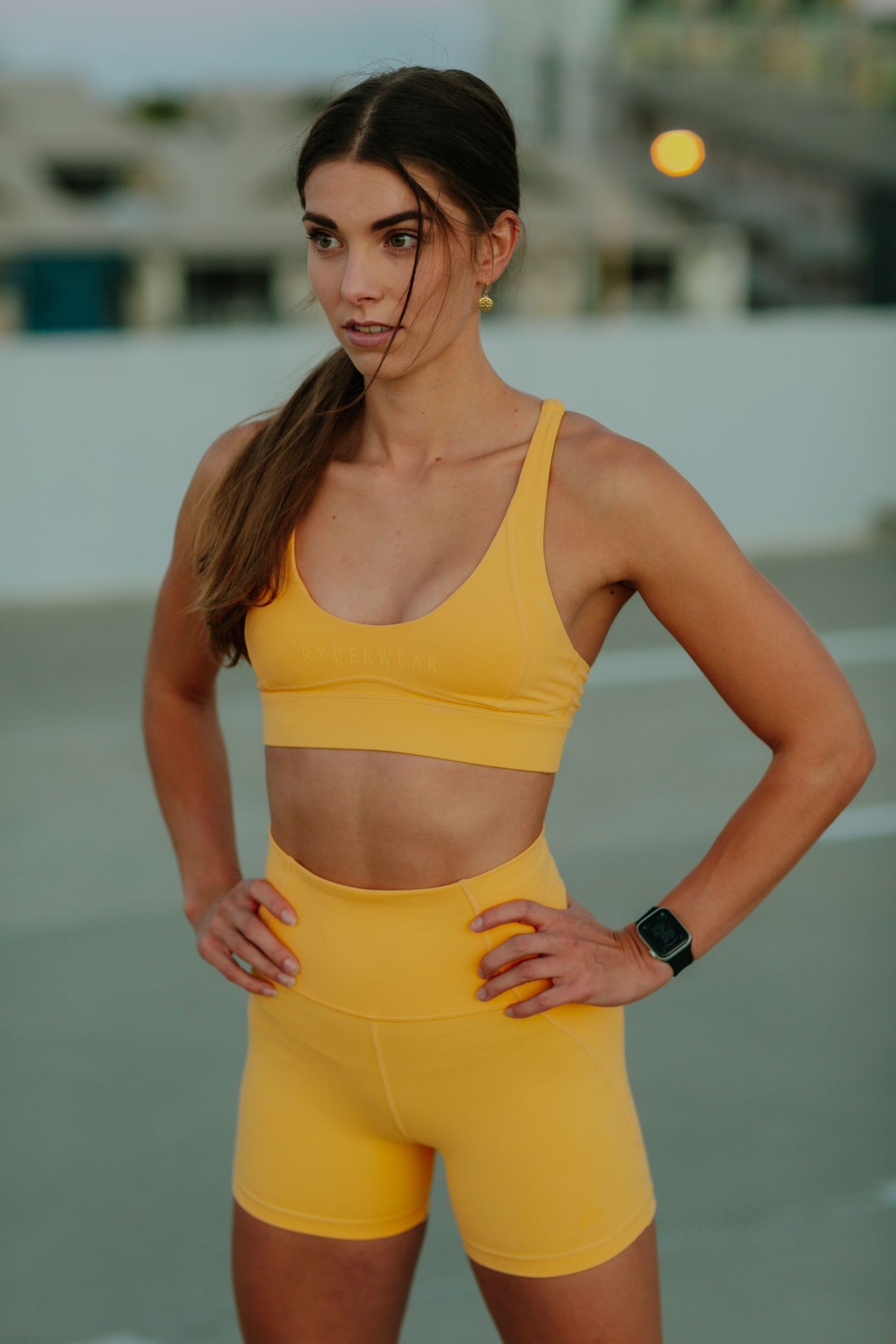 Athletic woman in yellow sports bra and shorts stands with hands on hips outdoors.