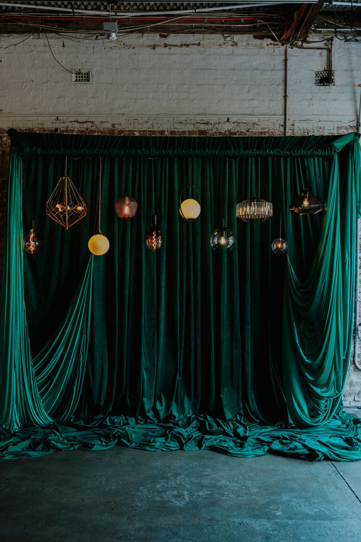 Eclectic mix of pendant lights hanging before draped emerald green velvet curtains in industrial space.
