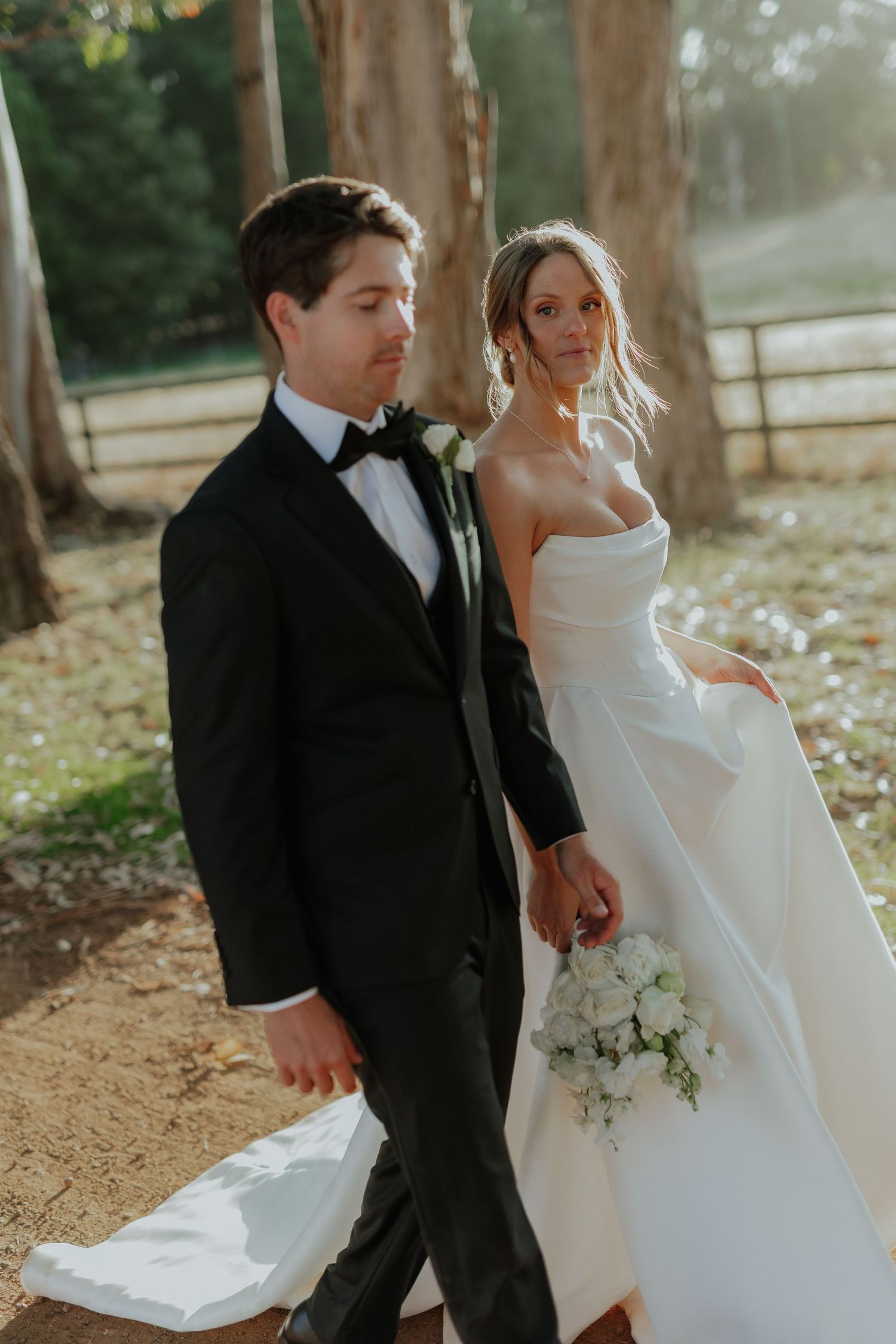 Bride and groom walking hand in hand through a sunlit forest, she holds a white bouquet.