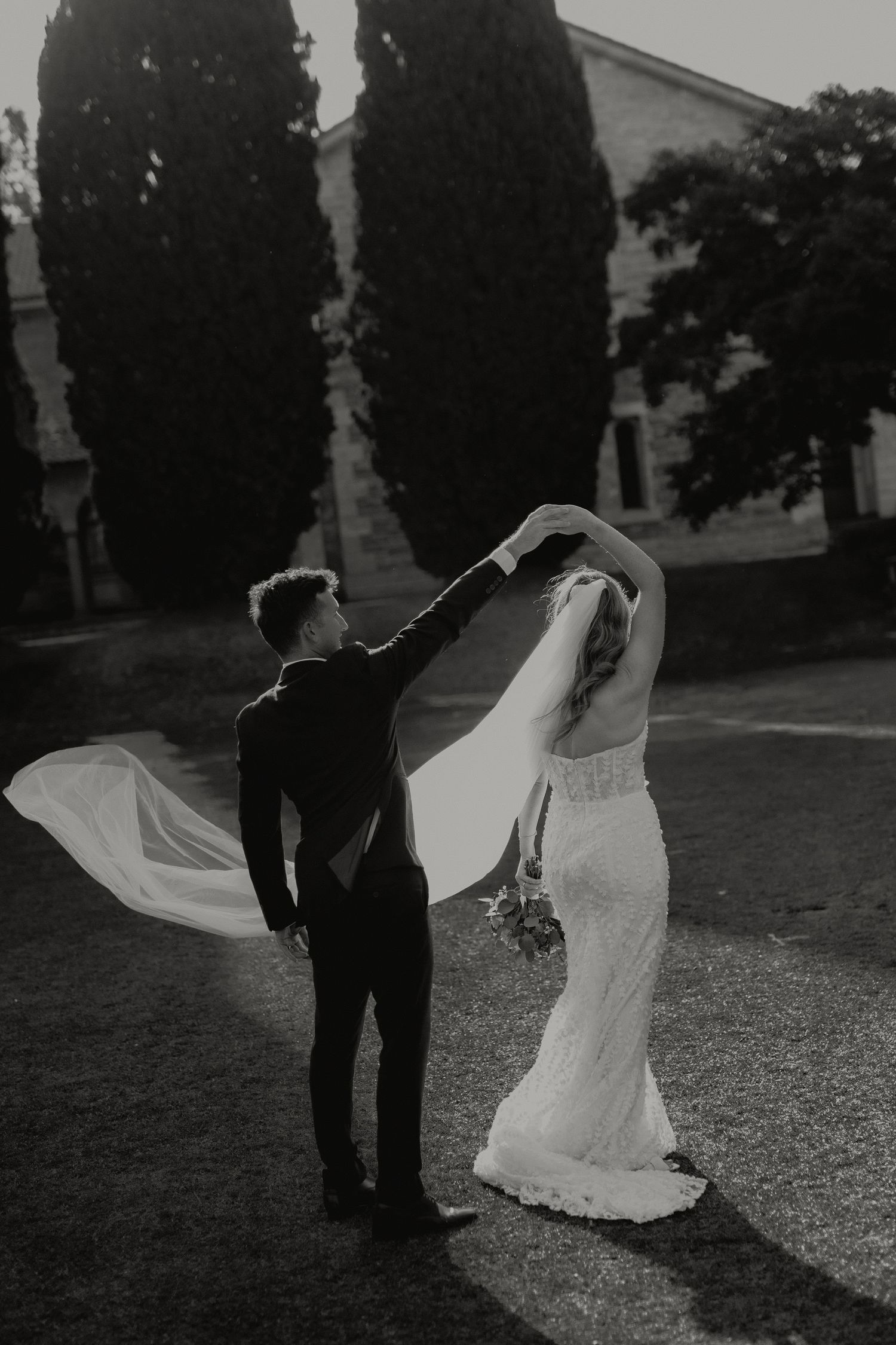 Black and white photo of bride and groom dancing outdoors, veil flowing in wind, cypress trees in background.
