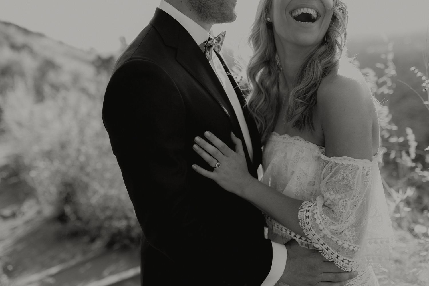 Black and white photo of laughing bride in off-shoulder gown embracing groom in tuxedo outdoors.