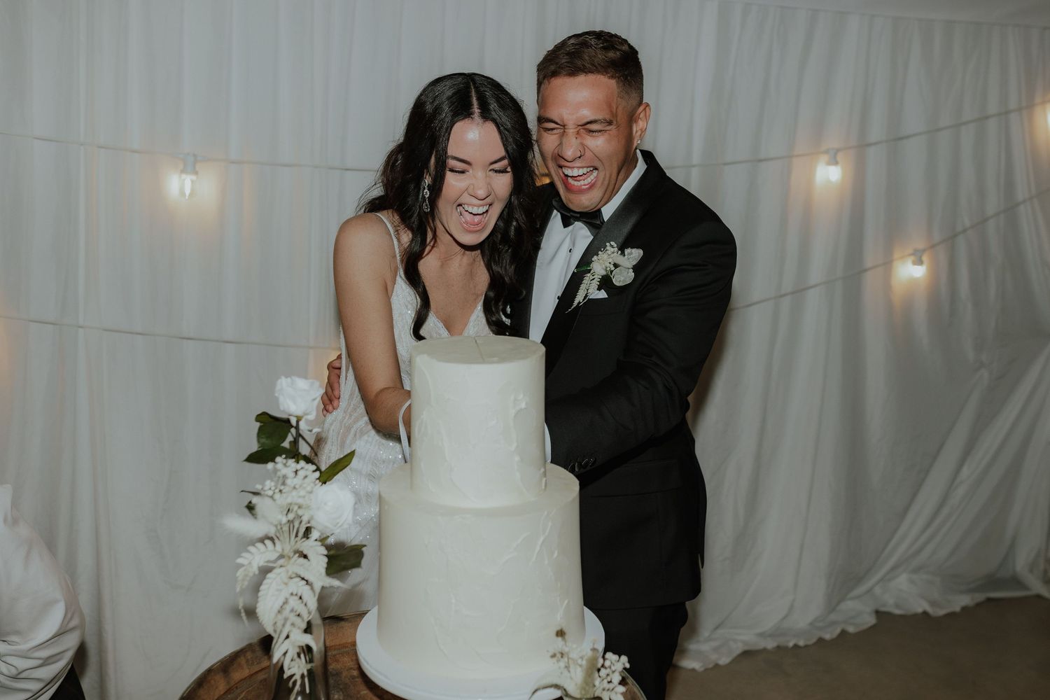 Smiling newlyweds cutting their elegant white three-tiered wedding cake at their reception.