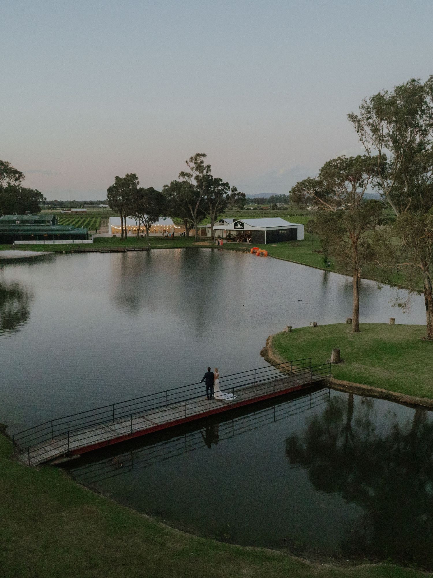 Aerial view of wedding venue with large pond, dock, and manicured grounds at sunset.