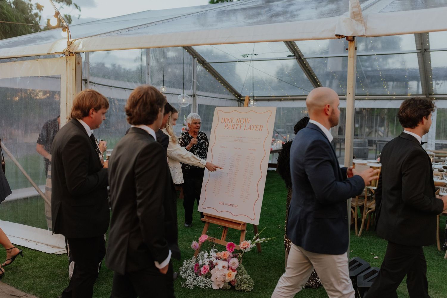 Wedding guests viewing seating chart on easel under clear marquee tent with floral arrangement.