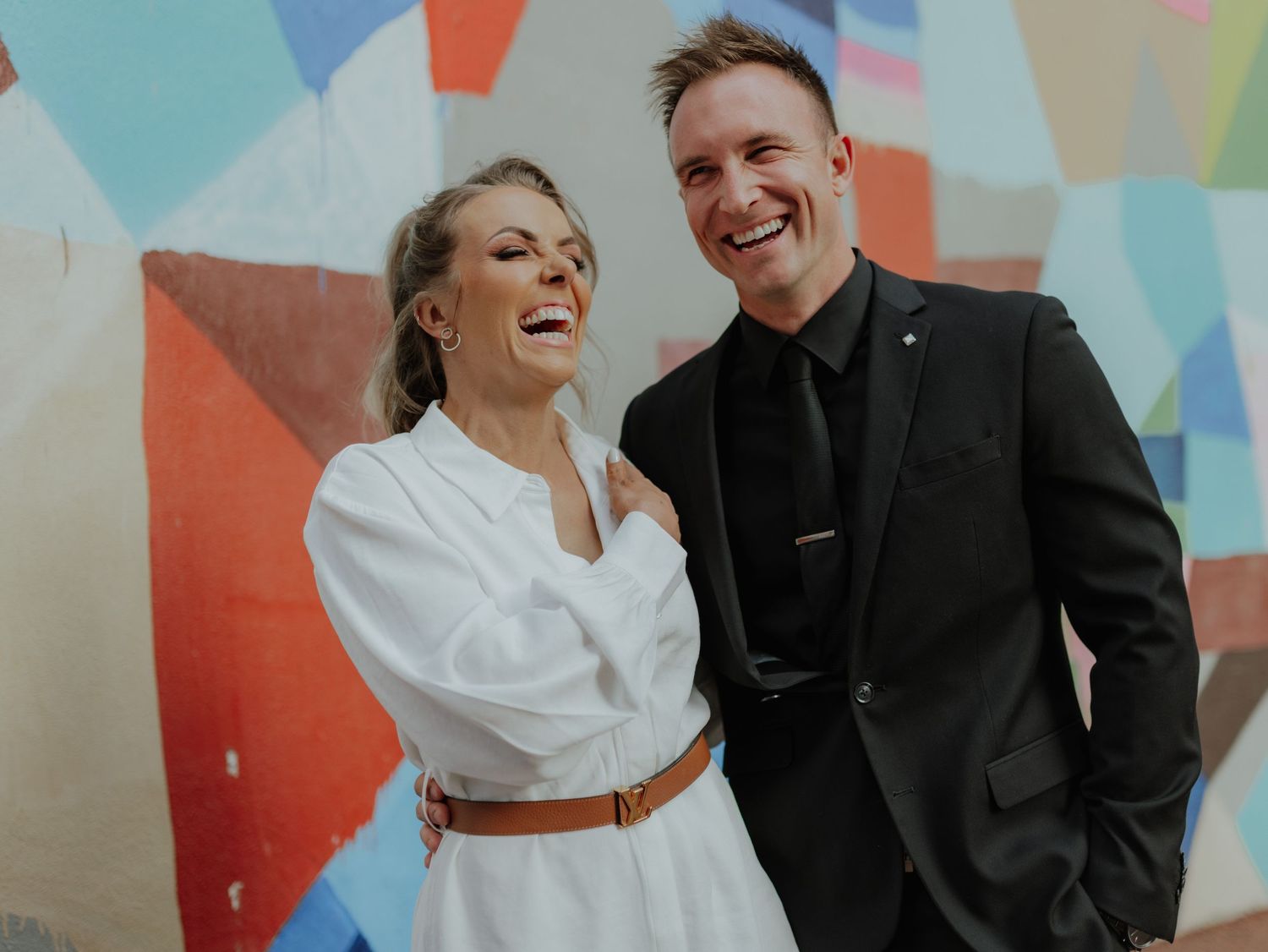 Laughing couple in white dress and black suit posing before colorful mural wall.
