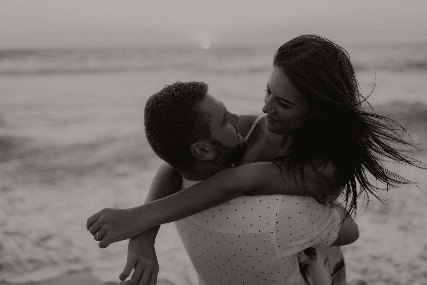Black and white photo of a couple embracing and laughing together on a beach at sunset.