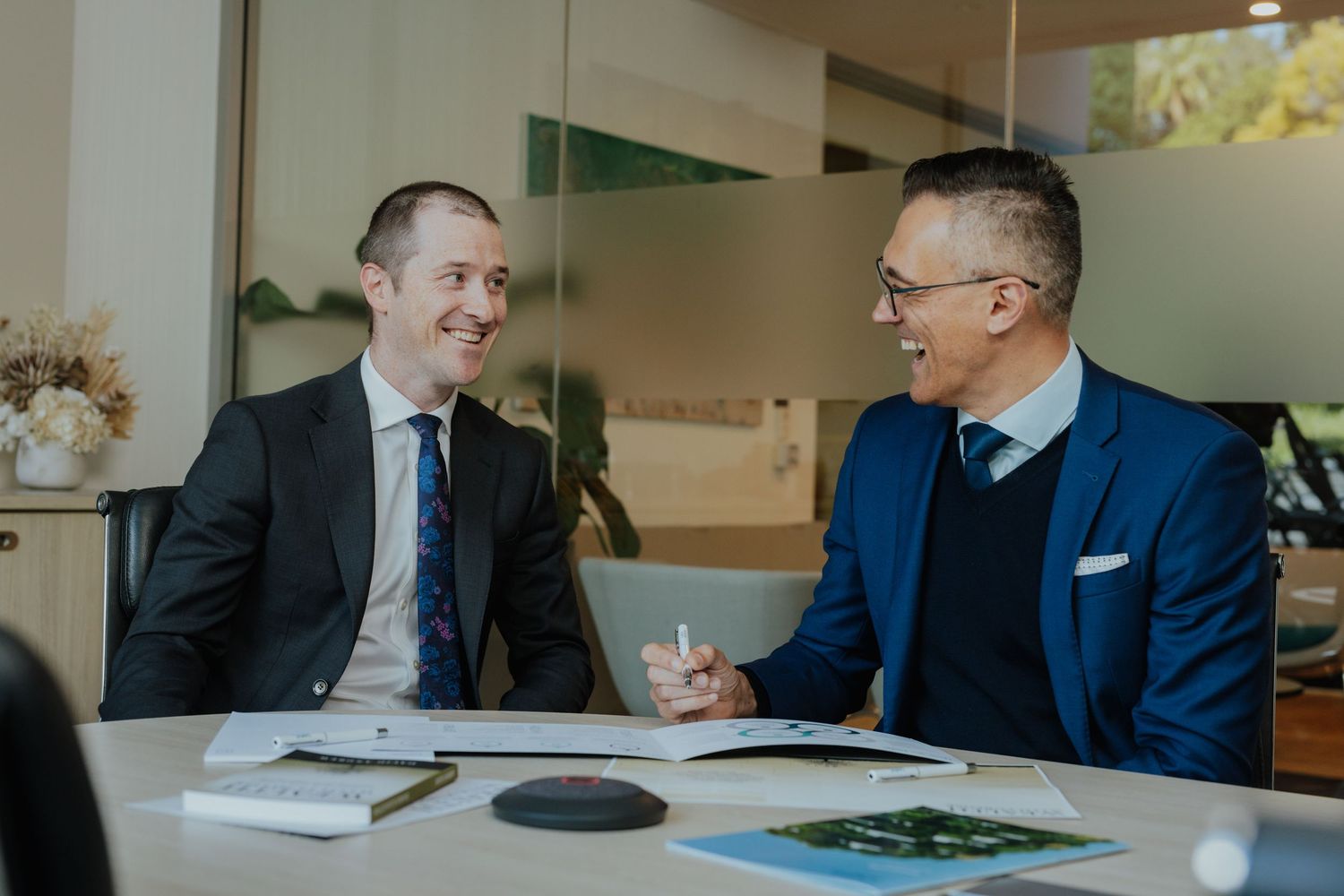 Two businessmen in suits smiling and shaking hands across a table in a bright modern office.