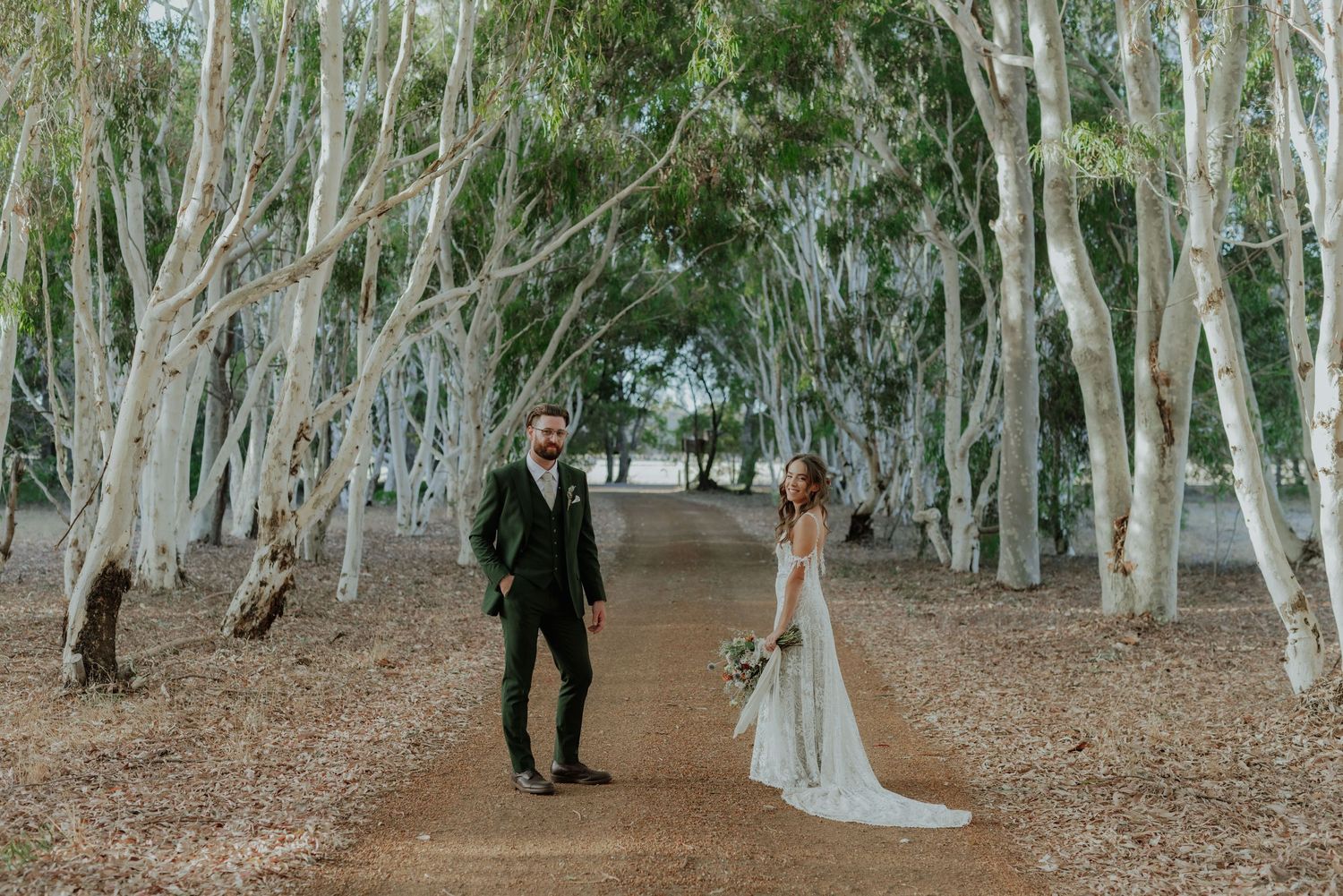 Bride and groom stand apart on dirt path lined with tall white-trunked trees in natural woodland setting.