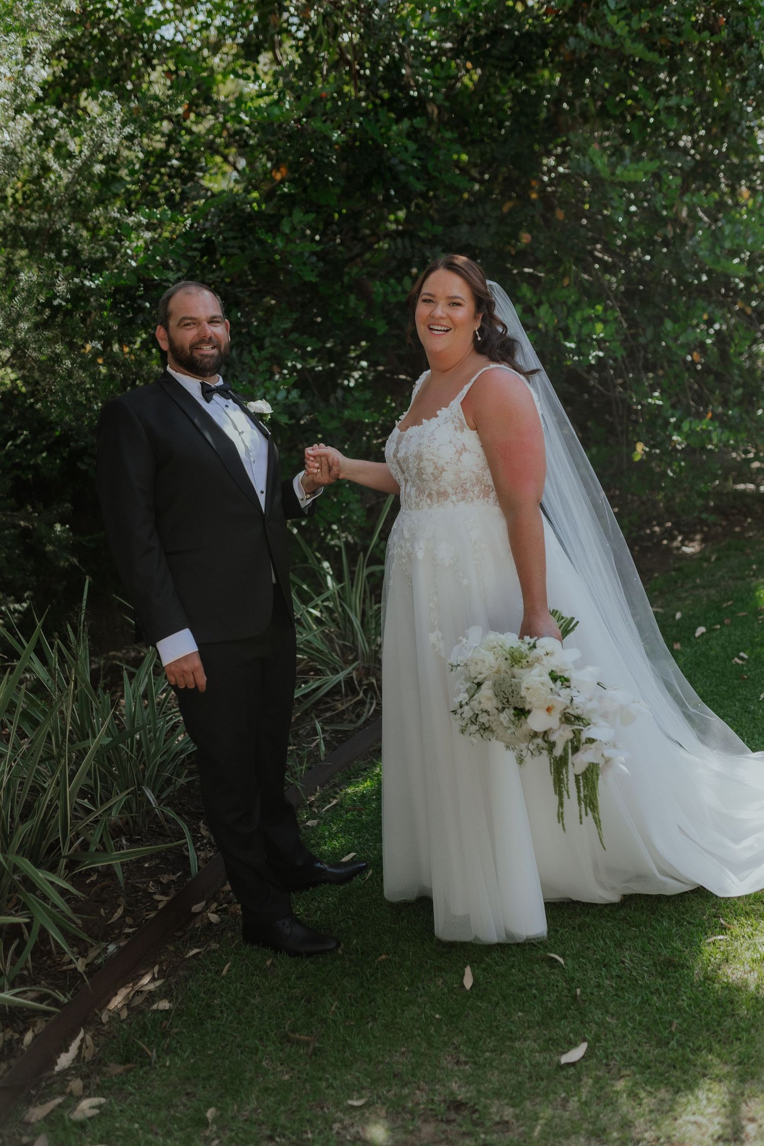 Bride and groom holding hands in lush garden, bride wearing white gown with veil and bouquet, groom in black tuxedo.