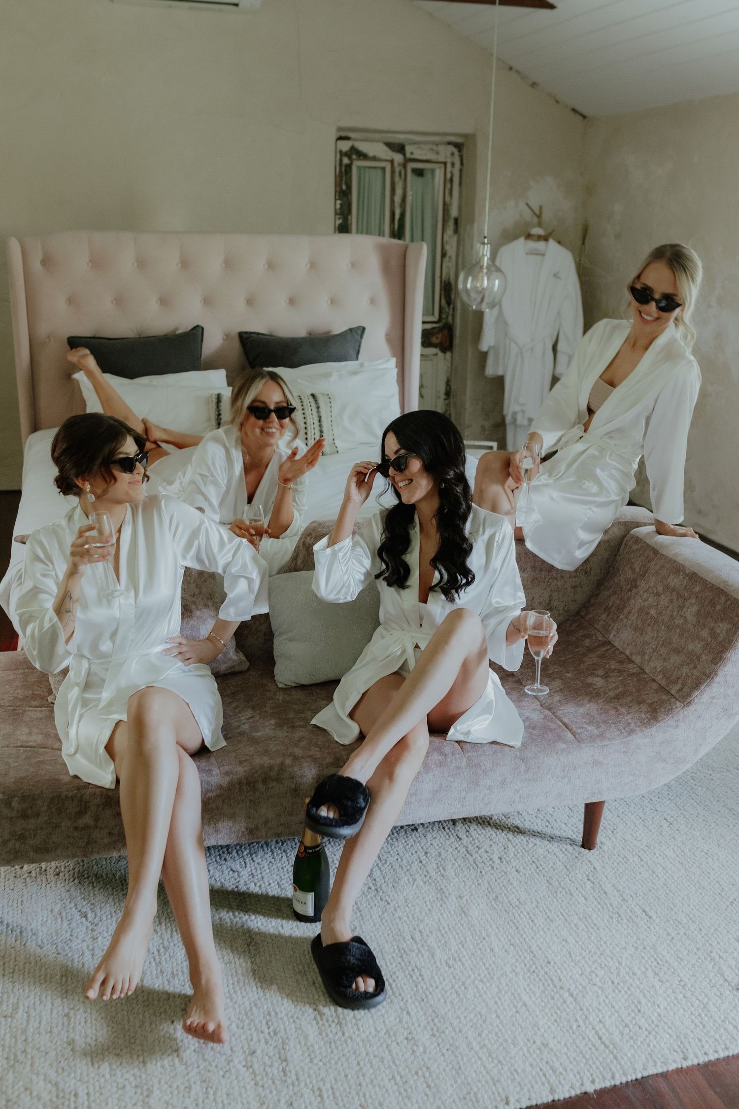 Bridesmaids in white robes and sunglasses celebrating with champagne on a velvet sofa before the wedding.