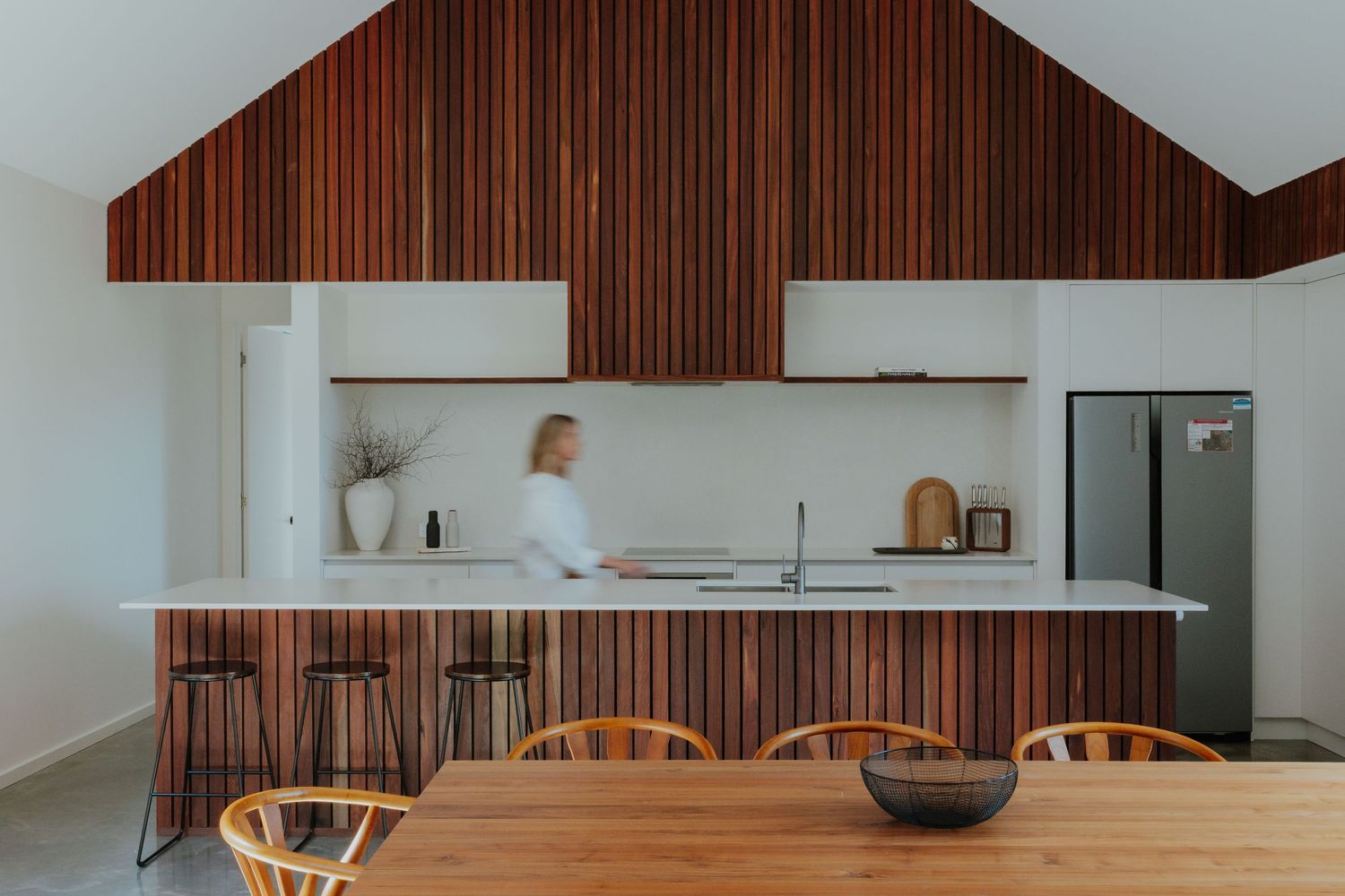 Open-plan kitchen with dramatic timber slat cathedral ceiling, white island bench and wooden dining furniture.